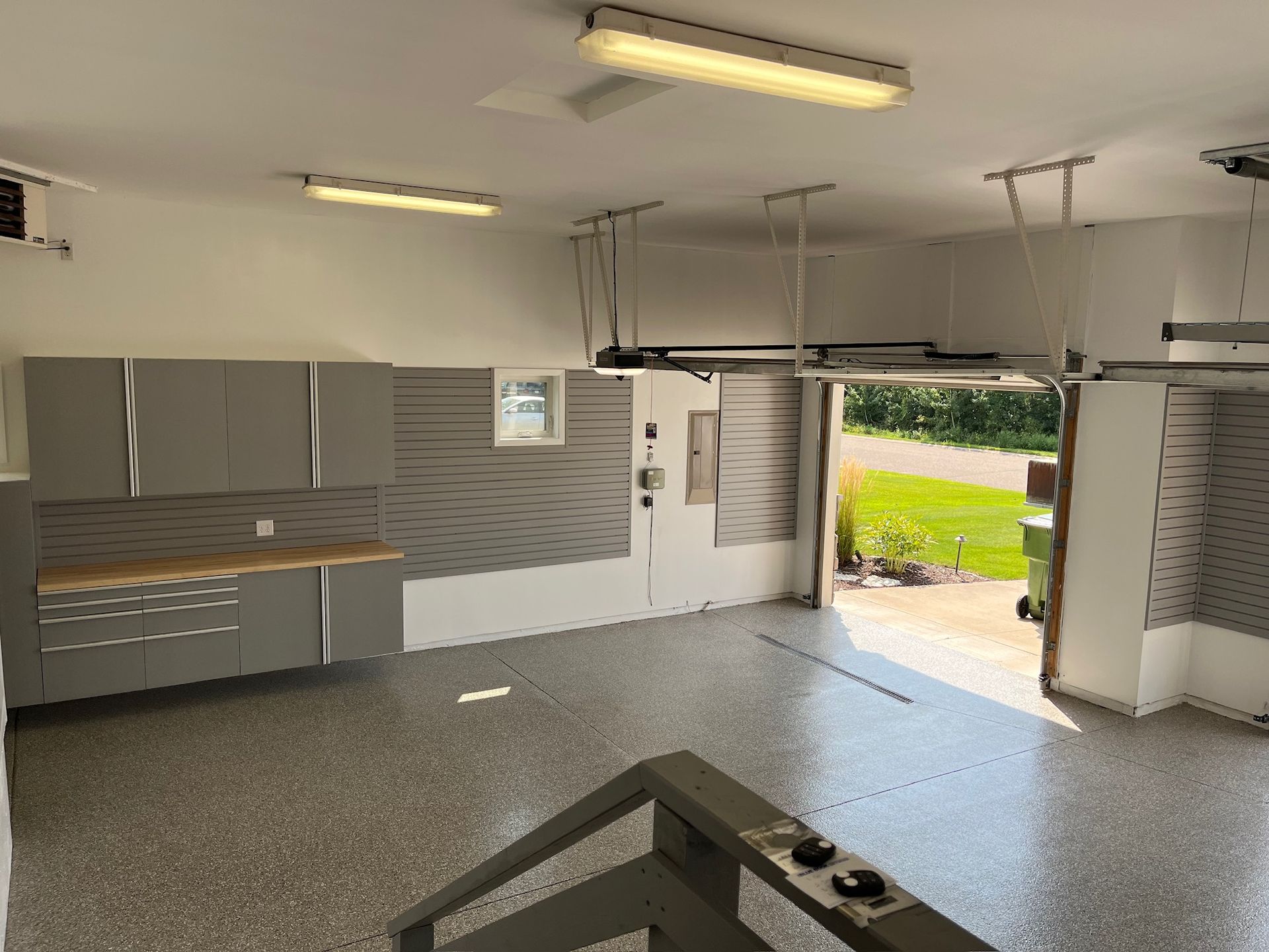 Empty garage with gray cabinets, speckled floor, open door, and bright daylight outside