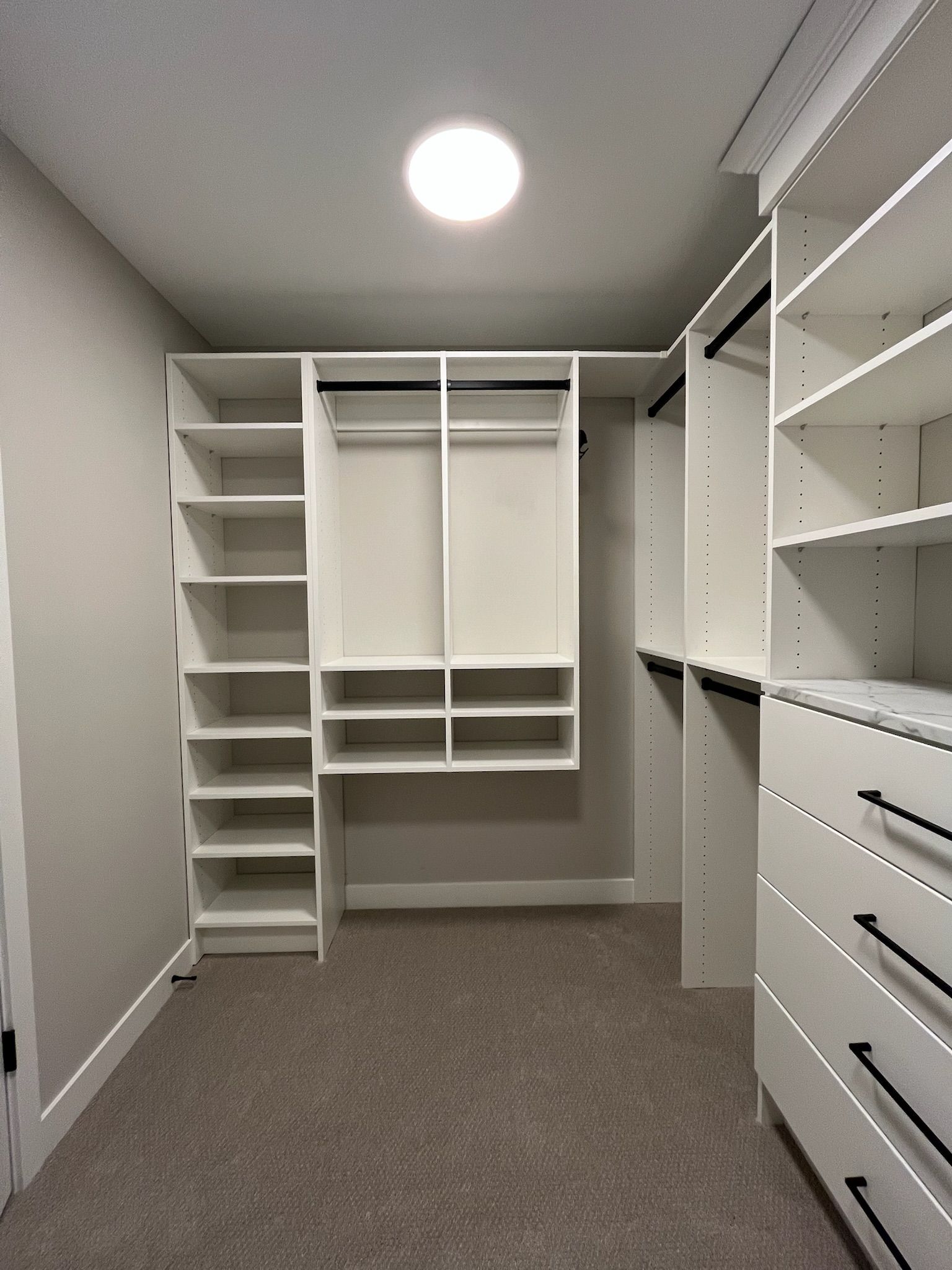 Empty walk-in closet with white shelves, hanging rods, drawers, and gray carpet.