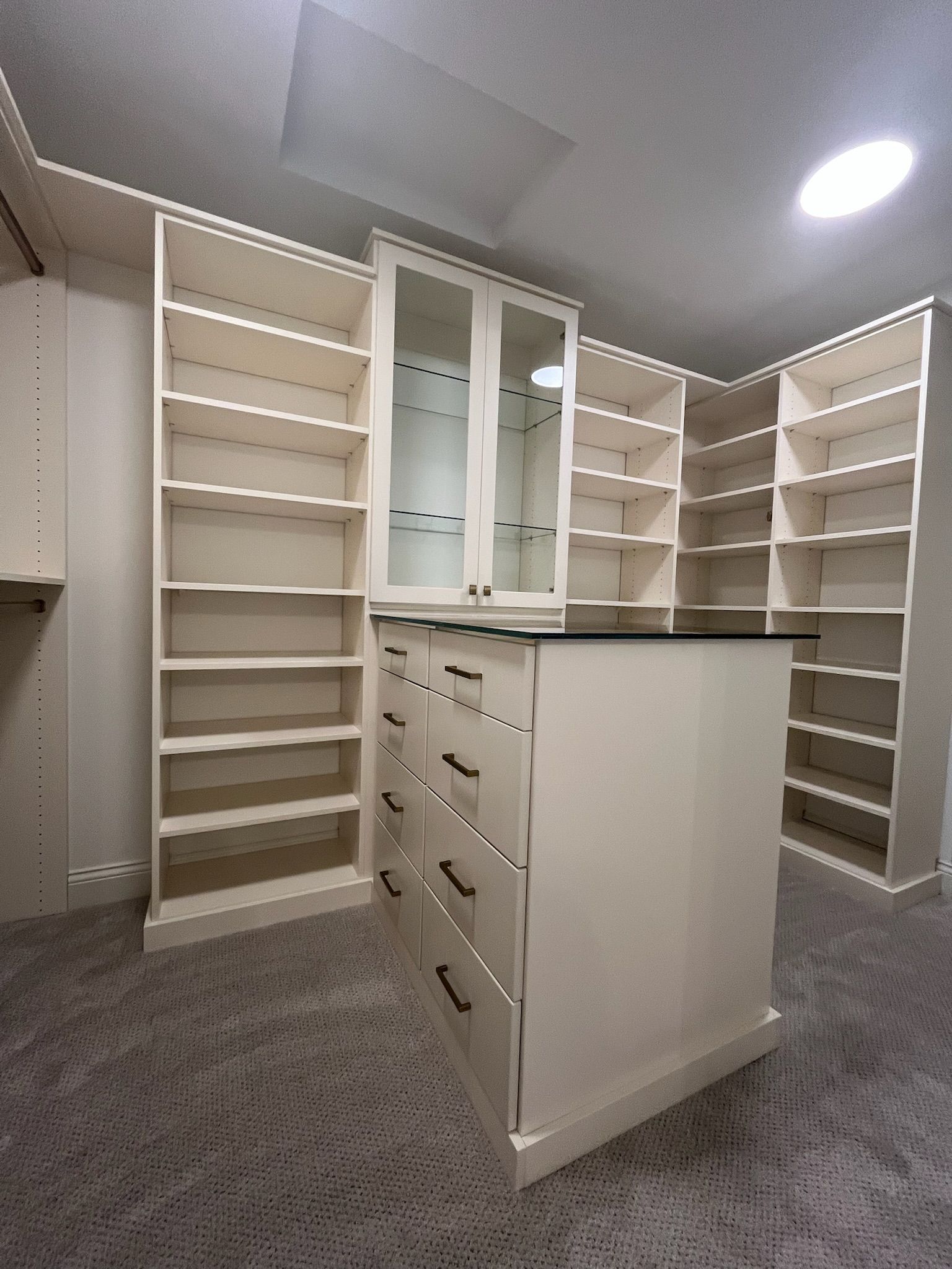 Empty walk-in closet with white shelves, drawers, and central island under ceiling lights.