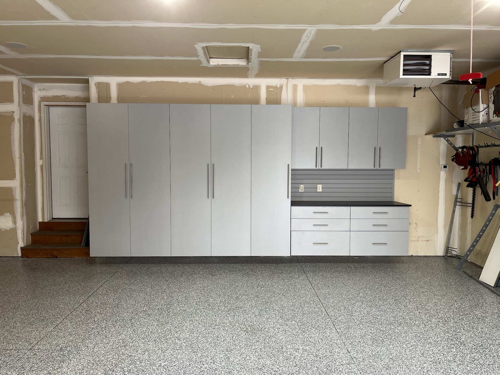 Garage with gray cabinets and workbench against unfinished drywall, with concrete floor and tools on the right.