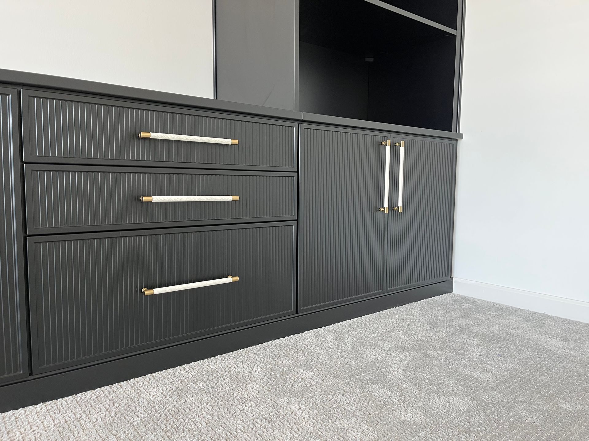 Gray ribbed media cabinet with gold handles on a light carpeted floor against a white wall.