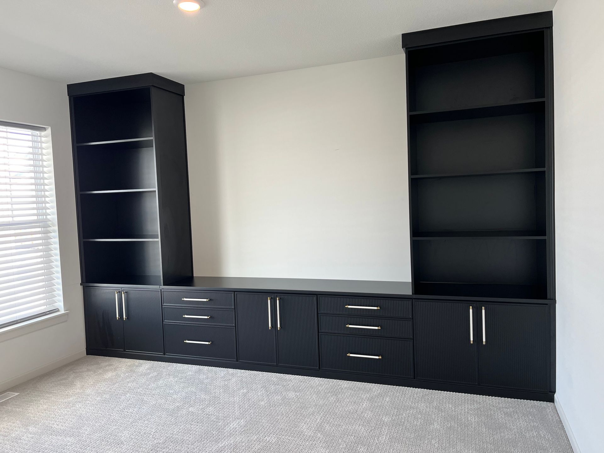 Black built-in cabinets and bookshelves along a wall in a carpeted room with a window on the left.