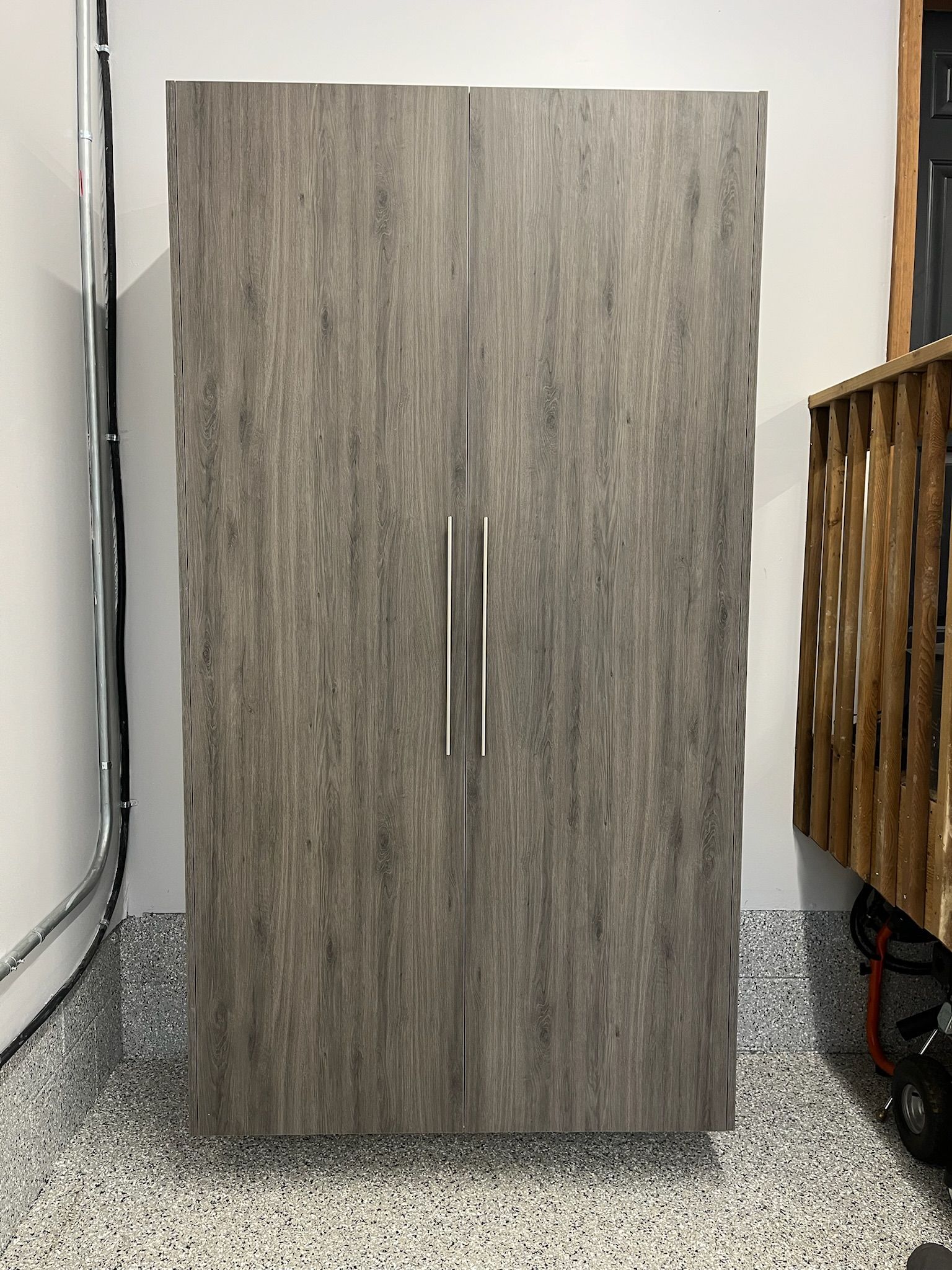 Gray wood-grain cabinet with two vertical silver handles in a room with a speckled floor and nearby furniture