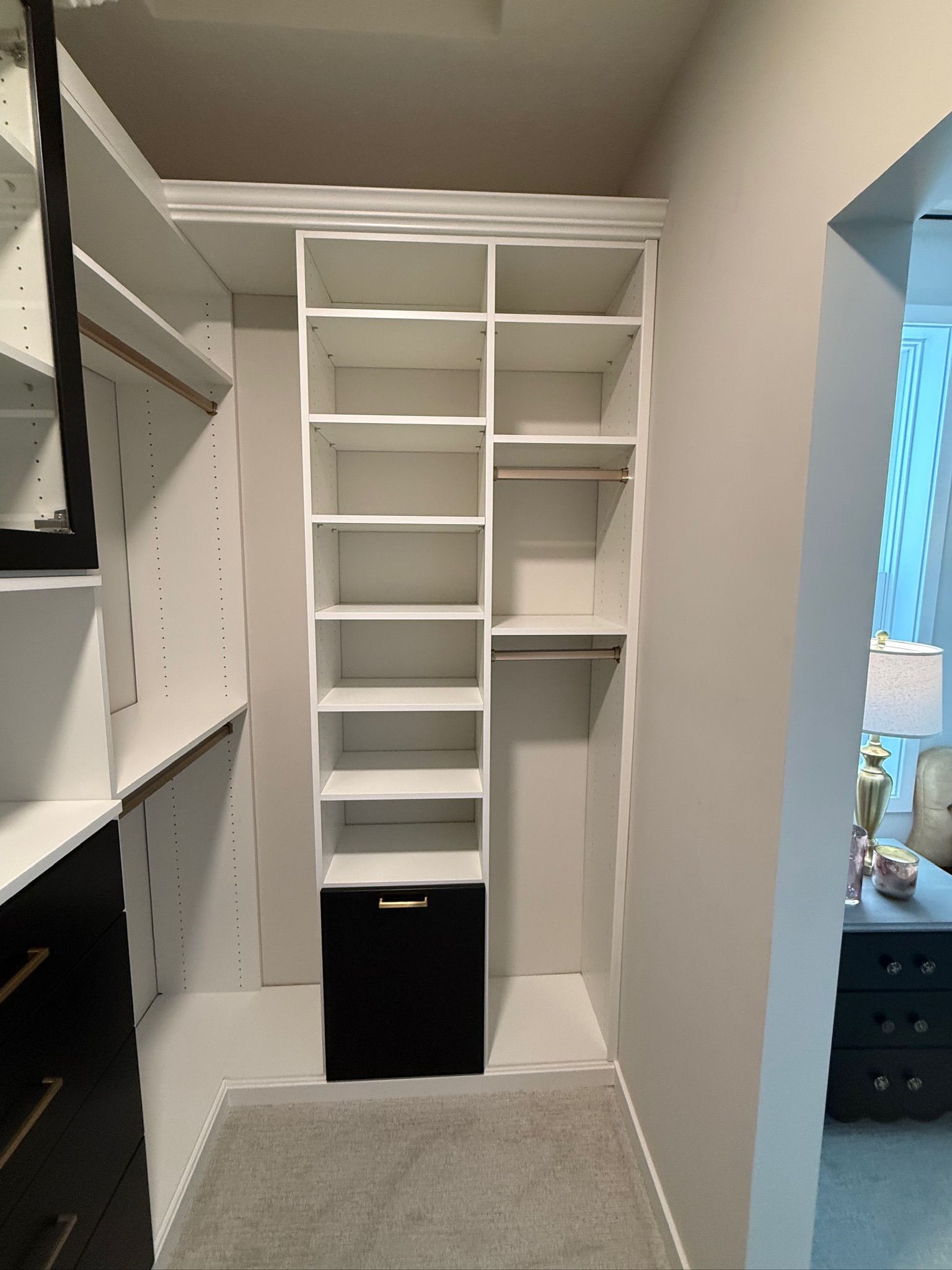 White built-in closet with empty shelves, hanging rods, and a black lower cabinet in a narrow room.