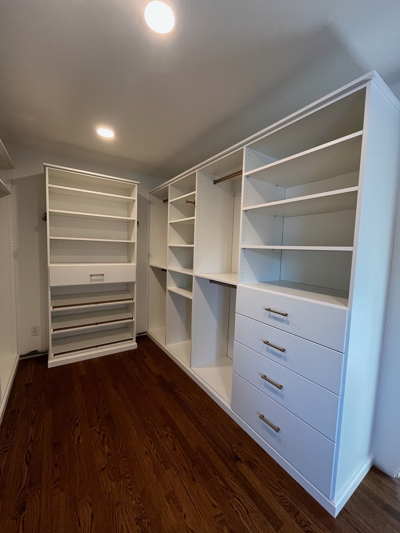 Empty walk-in closet with white built-in shelves and drawers, wood floor, and recessed ceiling lights.
