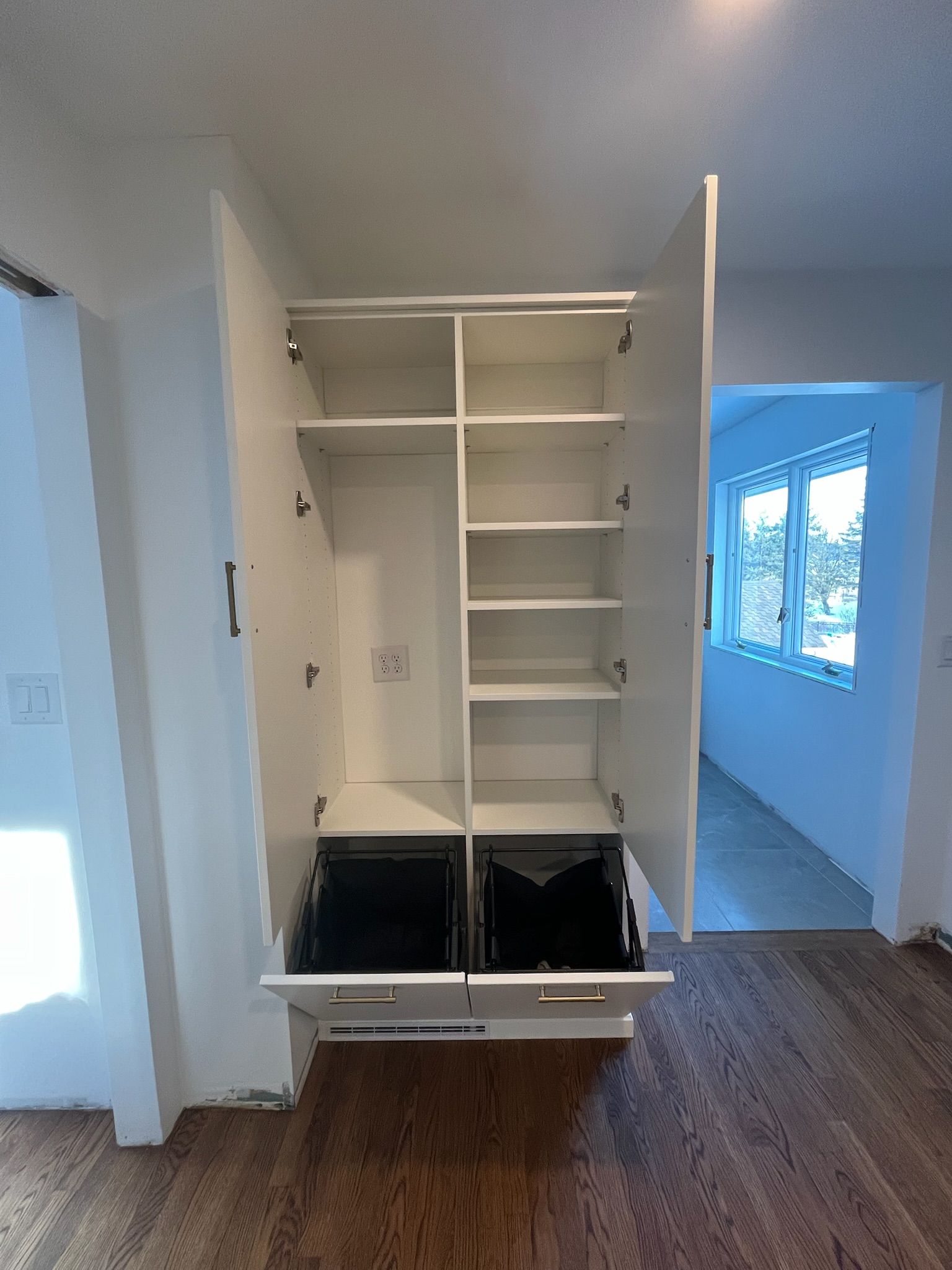 Open closet with shelves and two black bins in a hallway, beside a room with a window.