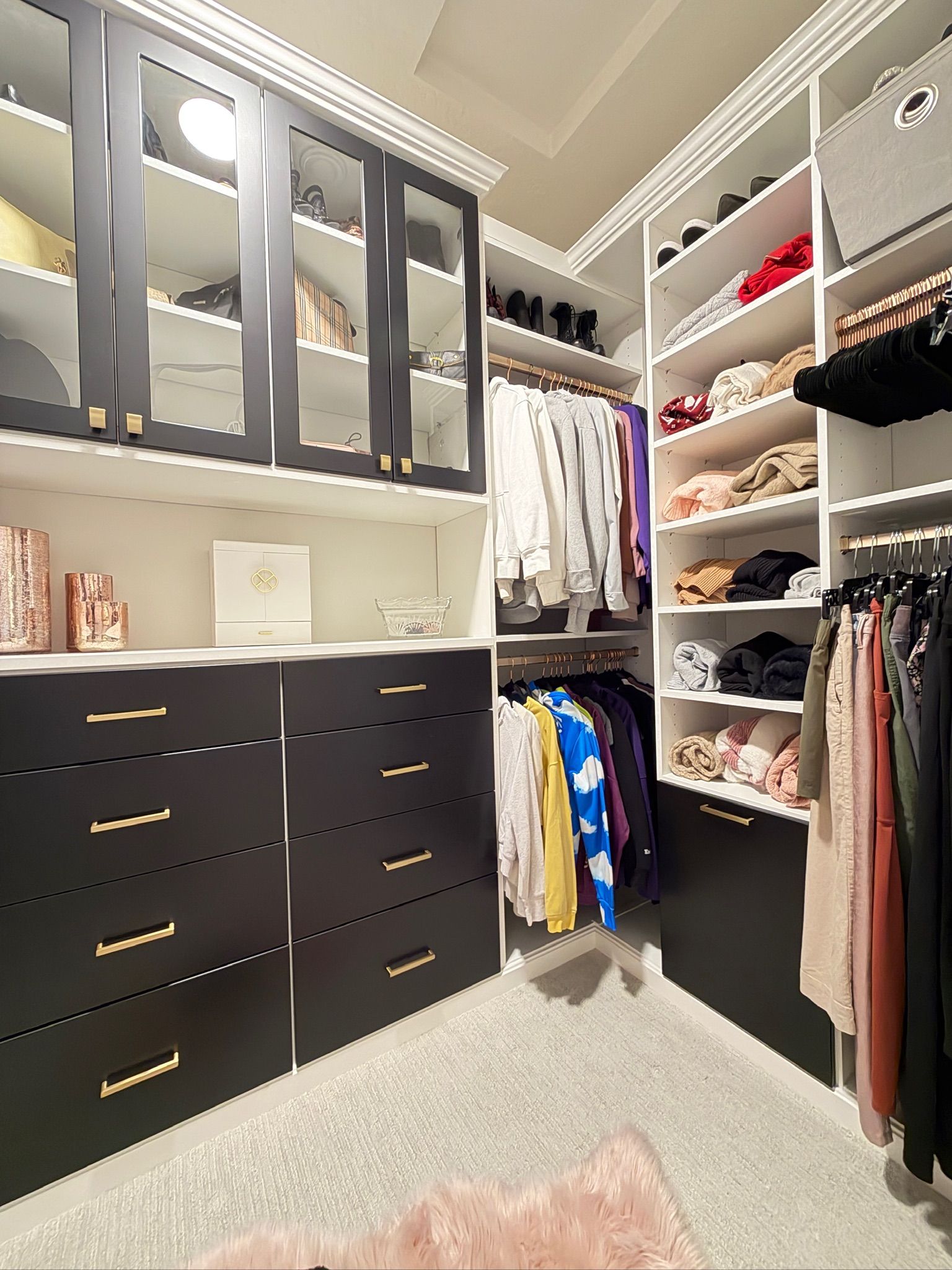 Walk-in closet with black drawers, white shelving, hanging clothes, and a pink rug