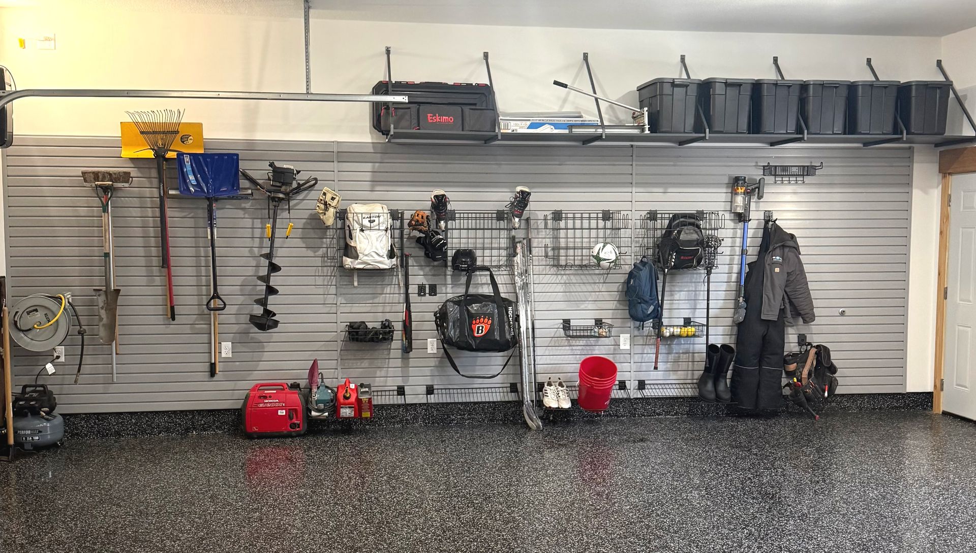 Garage wall with neatly organized tools, shelves, and red equipment on a slatwall above a speckled floor.