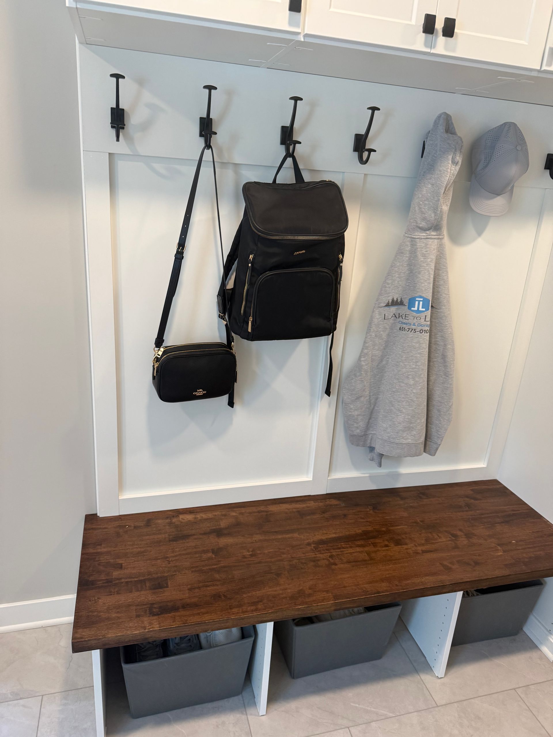 White entryway bench with black hooks holding bags and hats above gray storage bins below