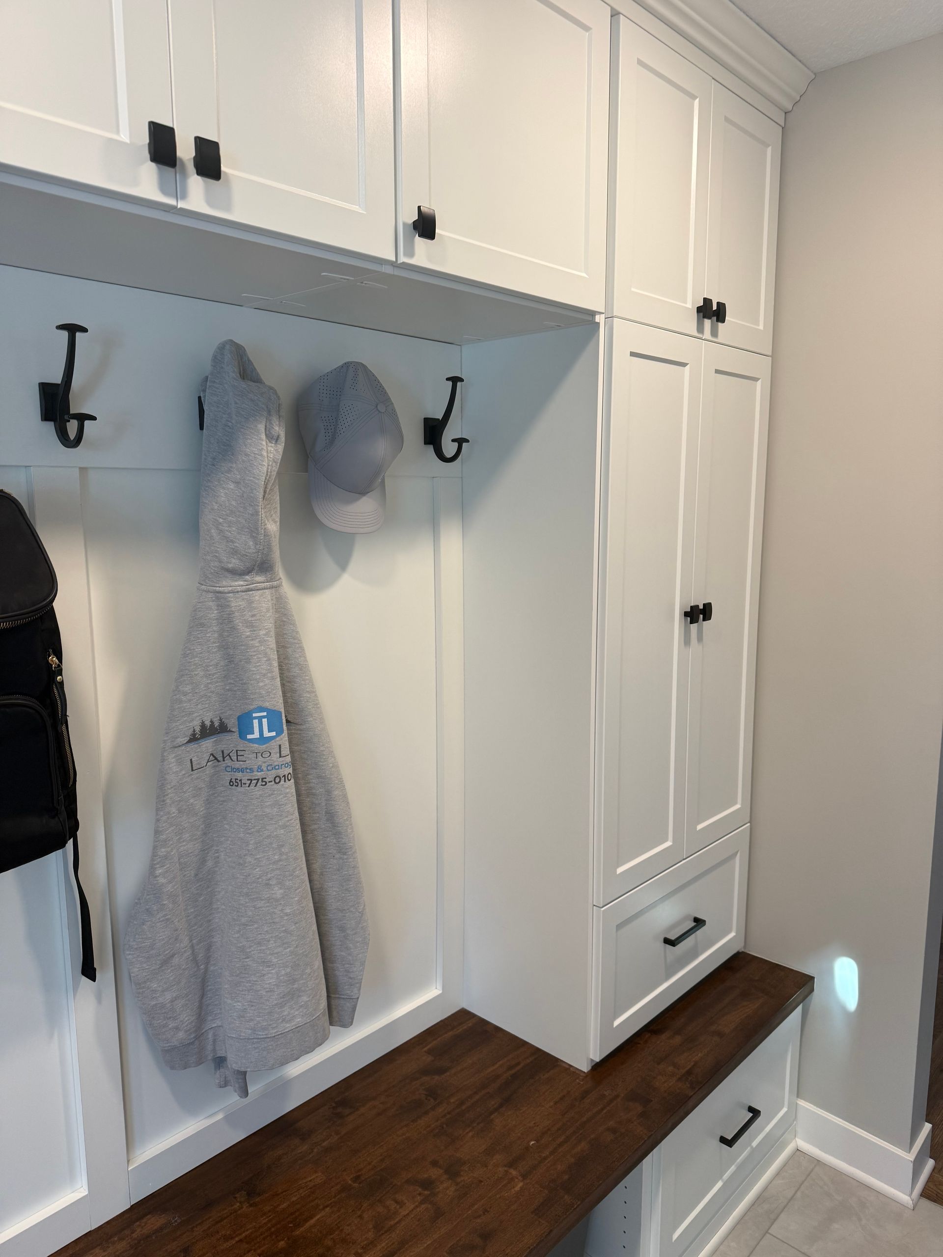 White built-in mudroom cabinets with hooks, coats, and a gray hat above a bench in a hallway.