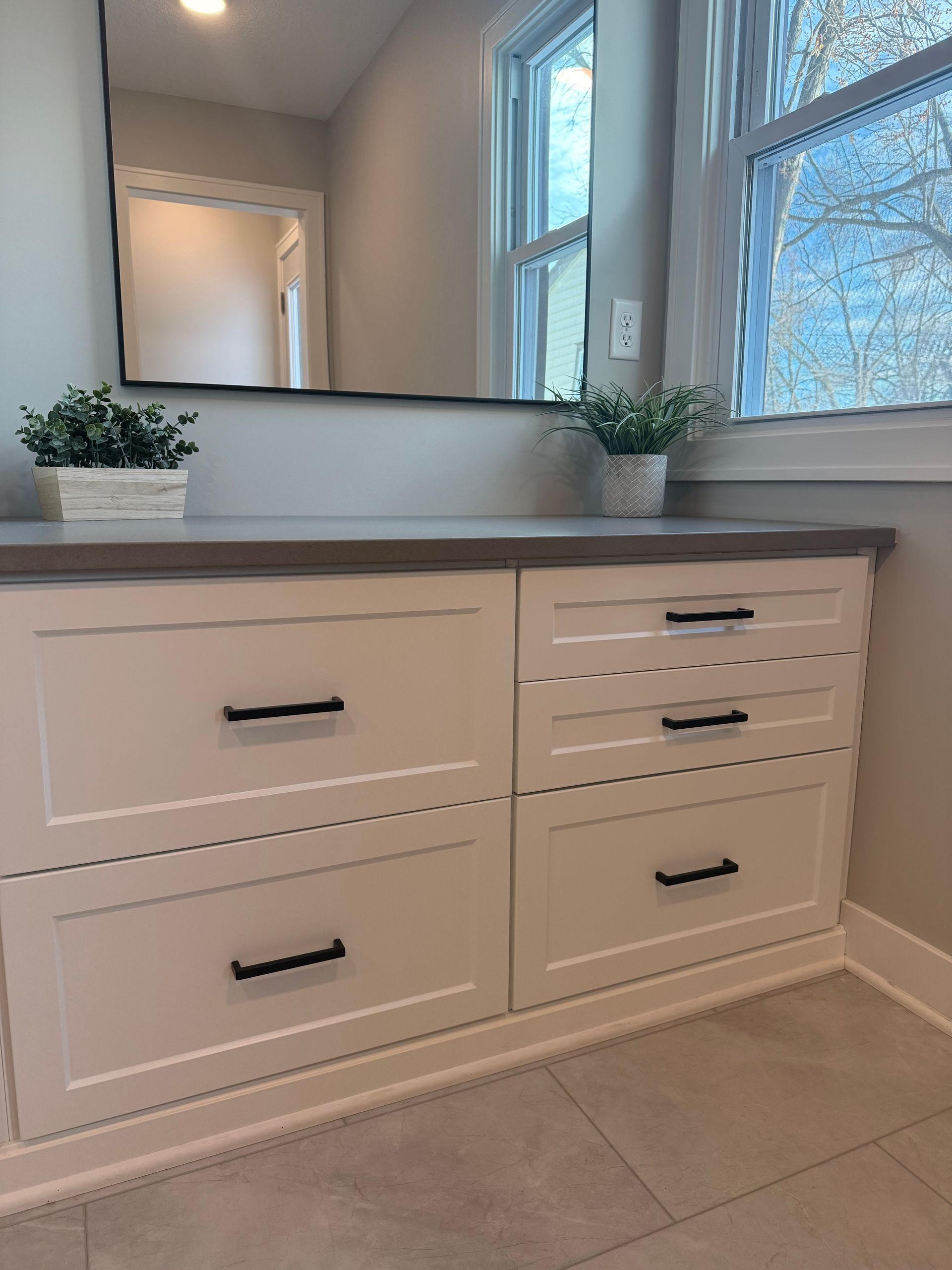 White bathroom vanity with drawers, double sink counter, mirror, plants, and a window with blinds.