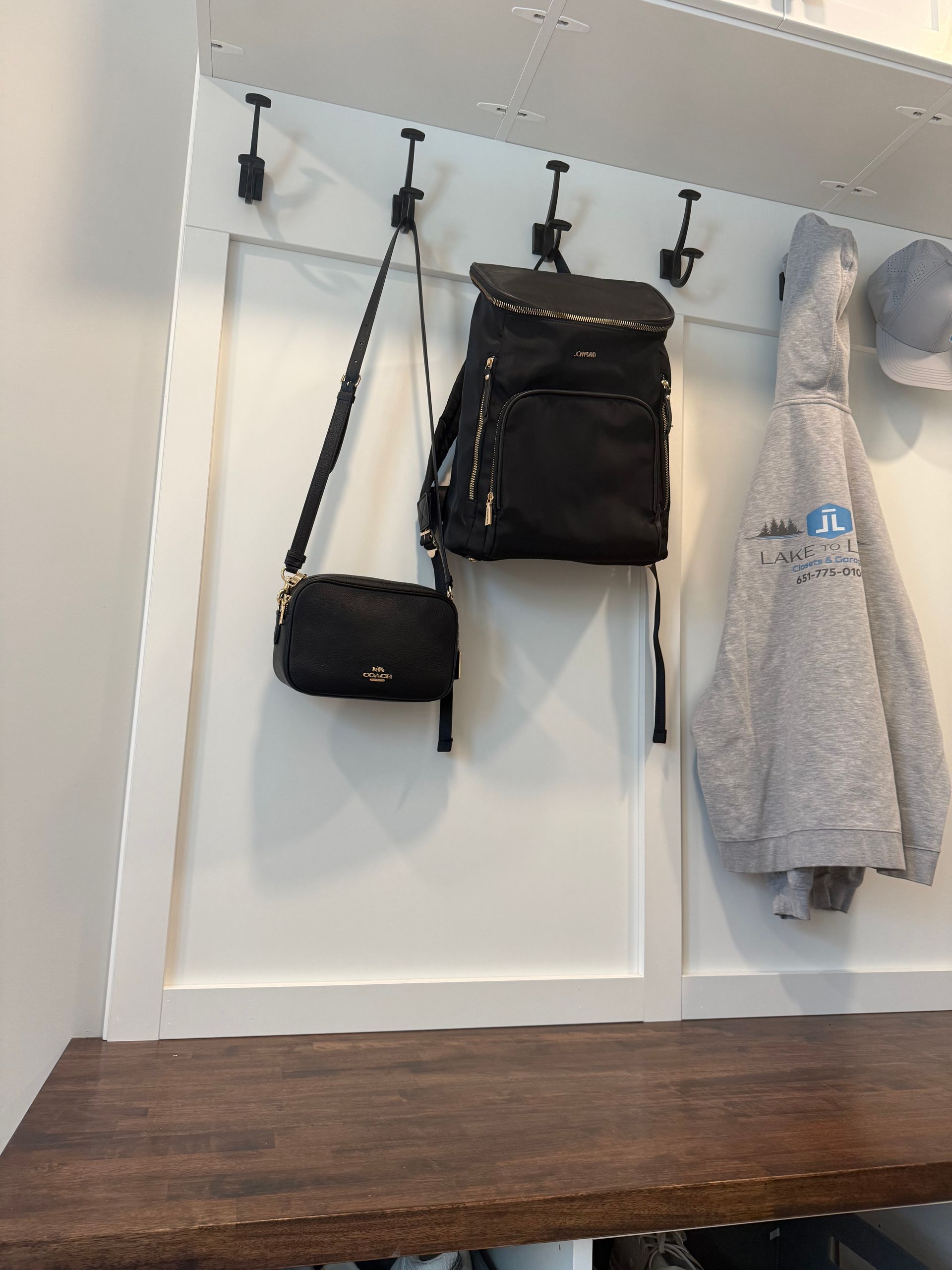 Wall hooks with black bag, crossbody purse, gray coat, and wooden bench in a white entryway