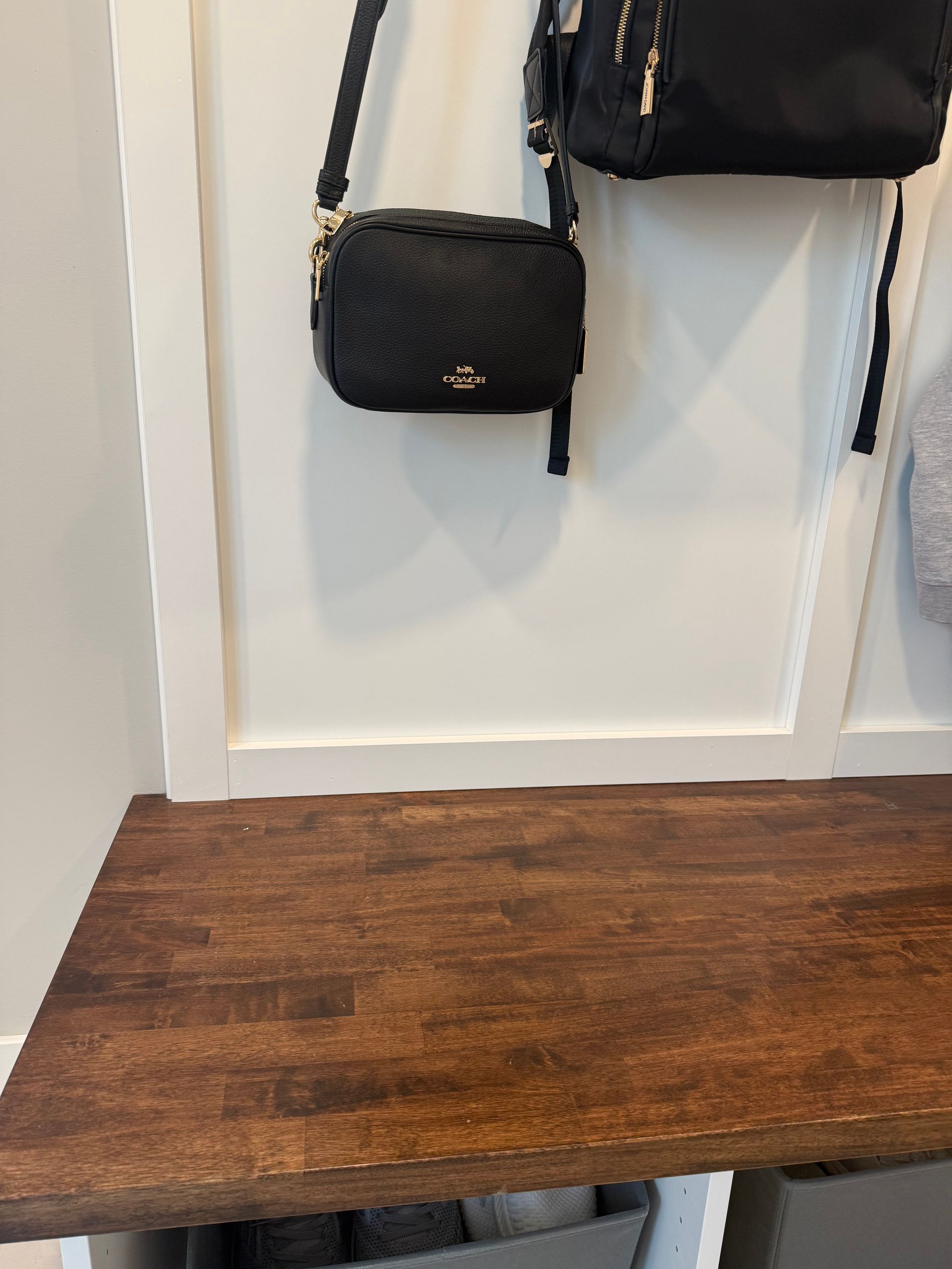 Wooden bench below hanging black handbags against a white wall.