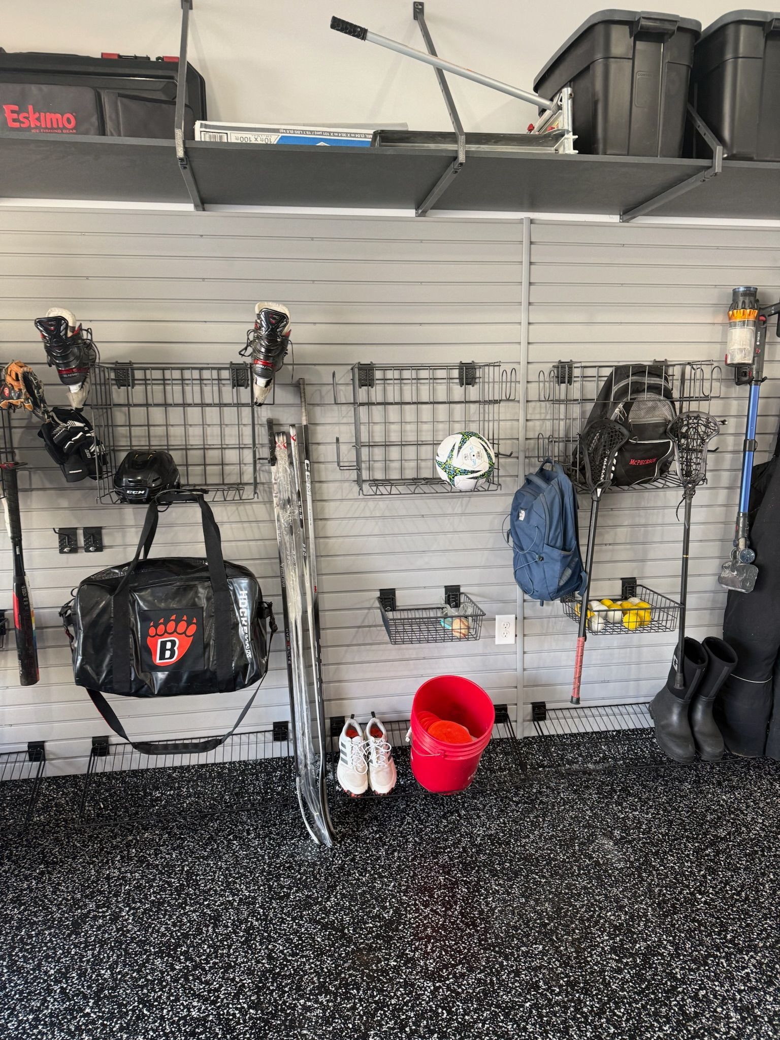 Garage wall with hanging tools, sports gear, storage bins, and a red bucket on a speckled floor