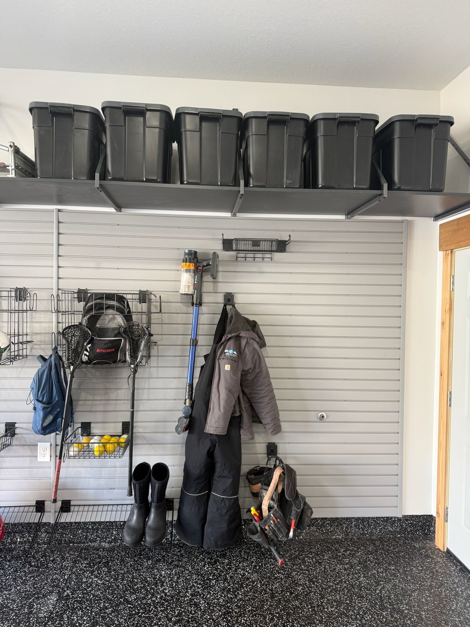 Garage wall with hanging coat, boots, and sports gear beneath a shelf of black storage bins.