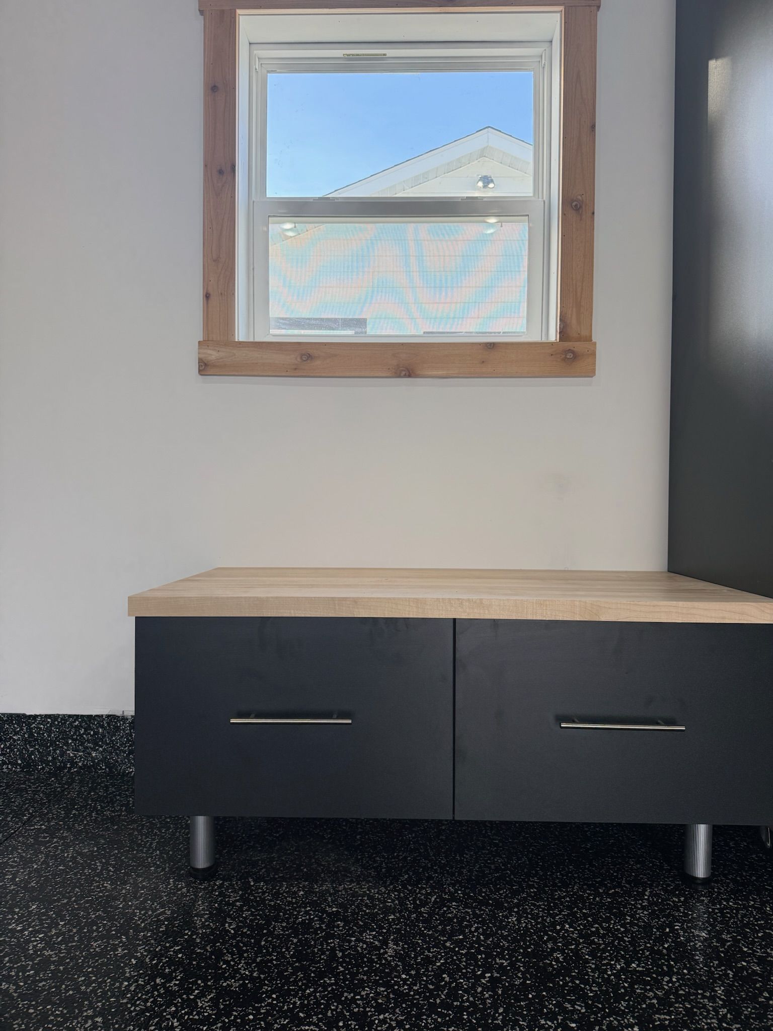 Small room with a wooden cabinet under a window, white walls, and black speckled flooring.