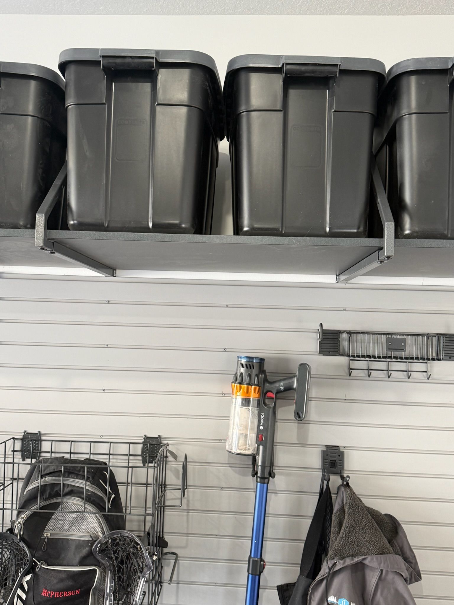 Garage wall with black storage bins on a shelf and tools hanging below on a white slatwall.