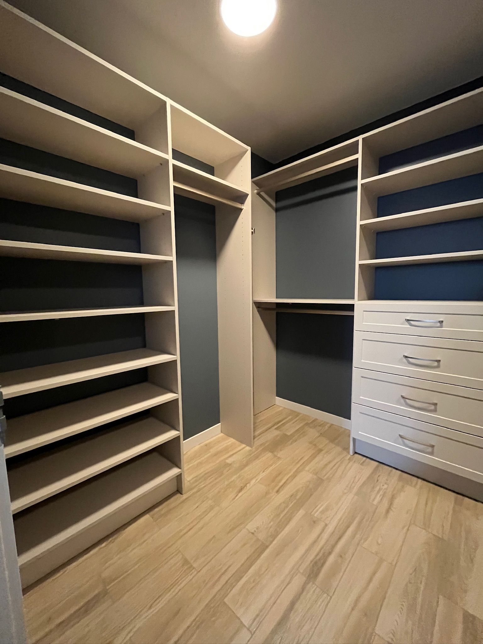 Walk-in closet with dark shelves, wood floor, and white drawer cabinets under ceiling light