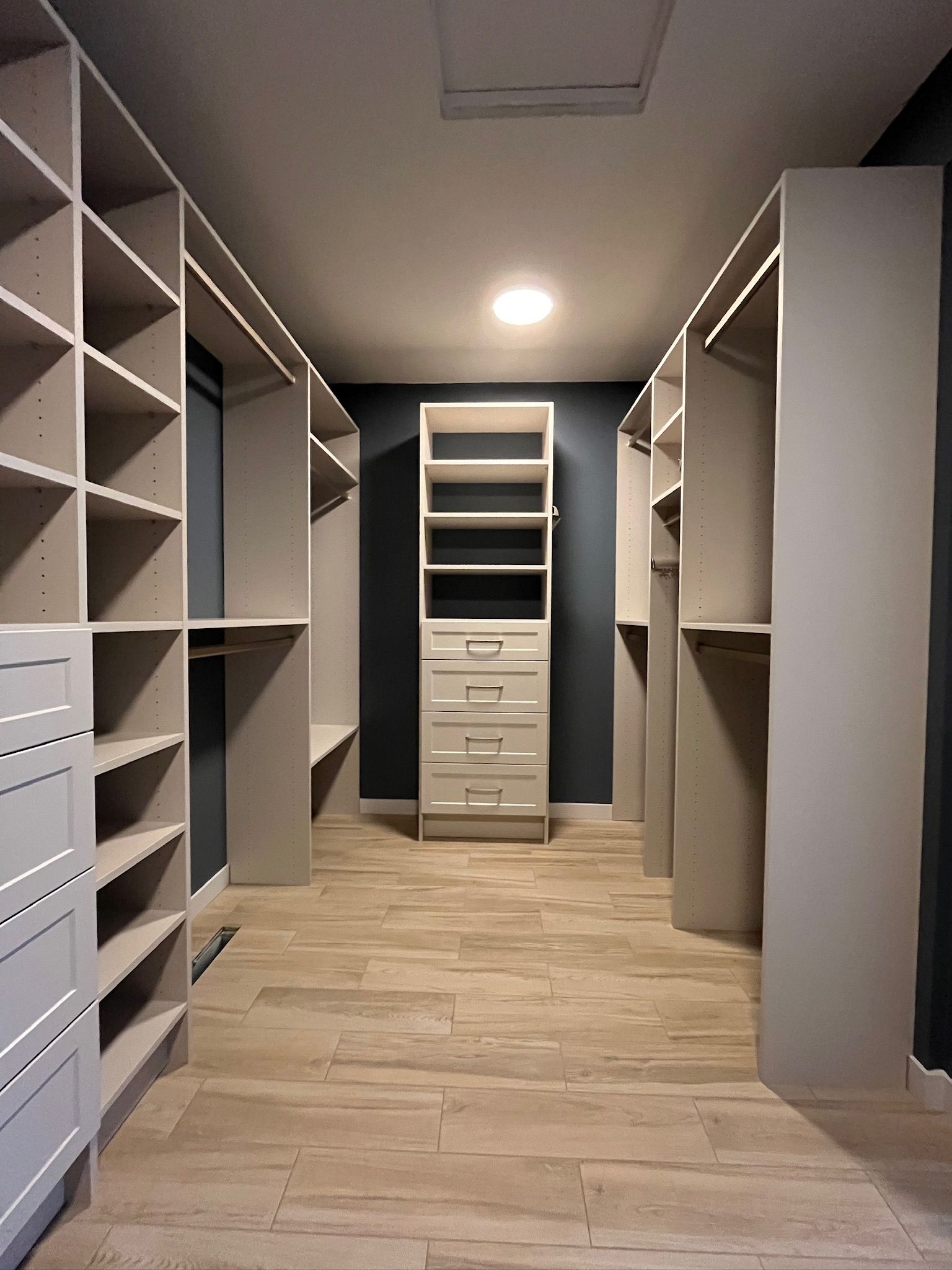 Walk-in closet with white shelving, drawers, and hanging spaces on both sides, wood-look floor and ceiling light.