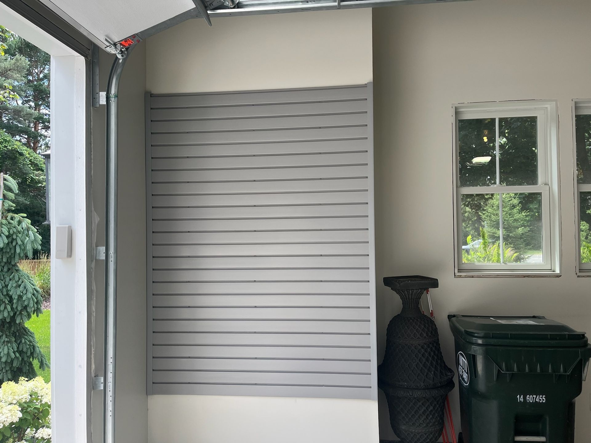 Garage opening with a closed gray slatted door, white wall, and outdoor items including a vase and trash bin.