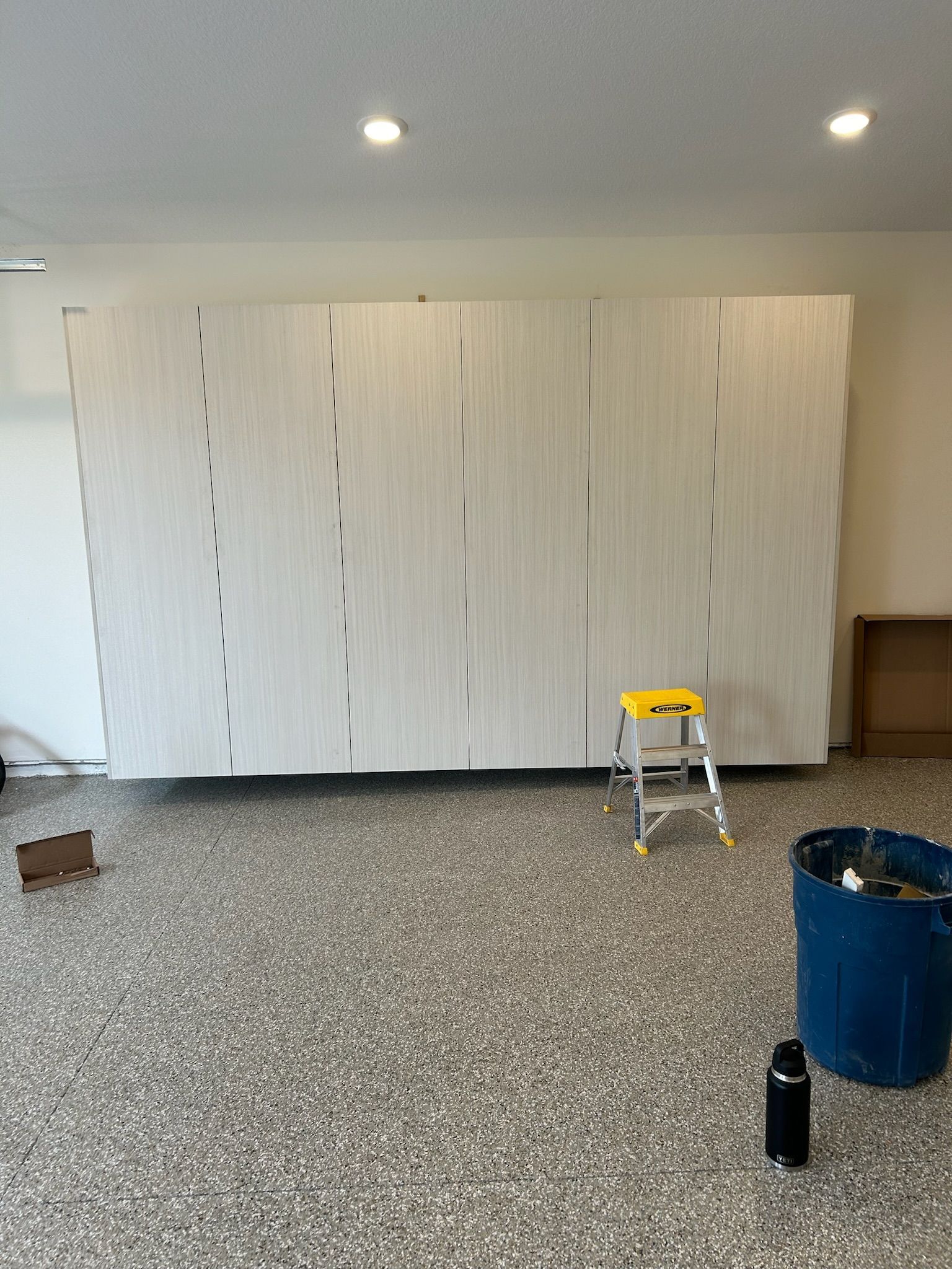 Large white cabinets in a room under renovation, with a step stool and cleaning supplies on a speckled floor.