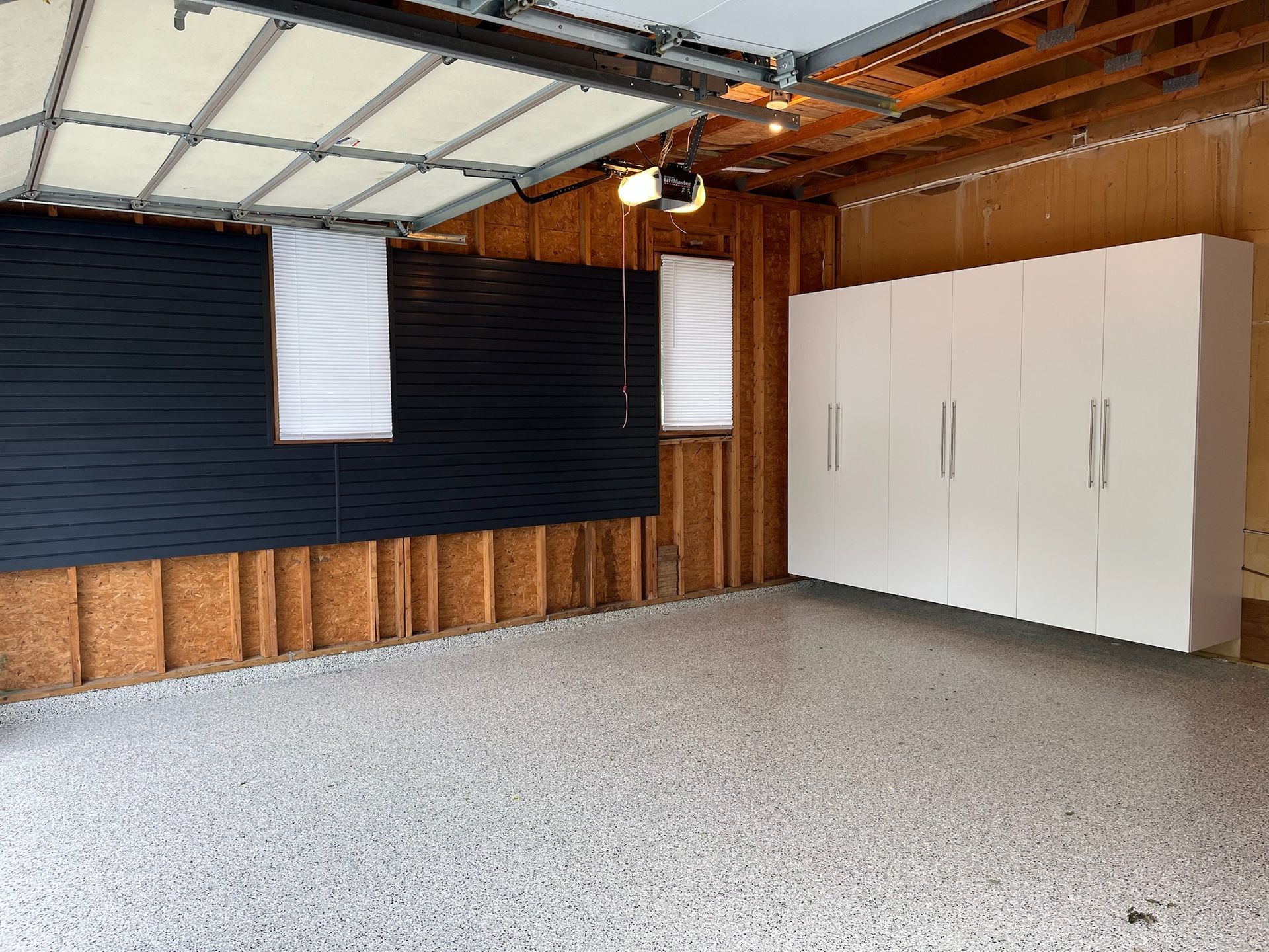 Empty garage with speckled floor, black wall panels, white cabinets, and hanging lights and shades