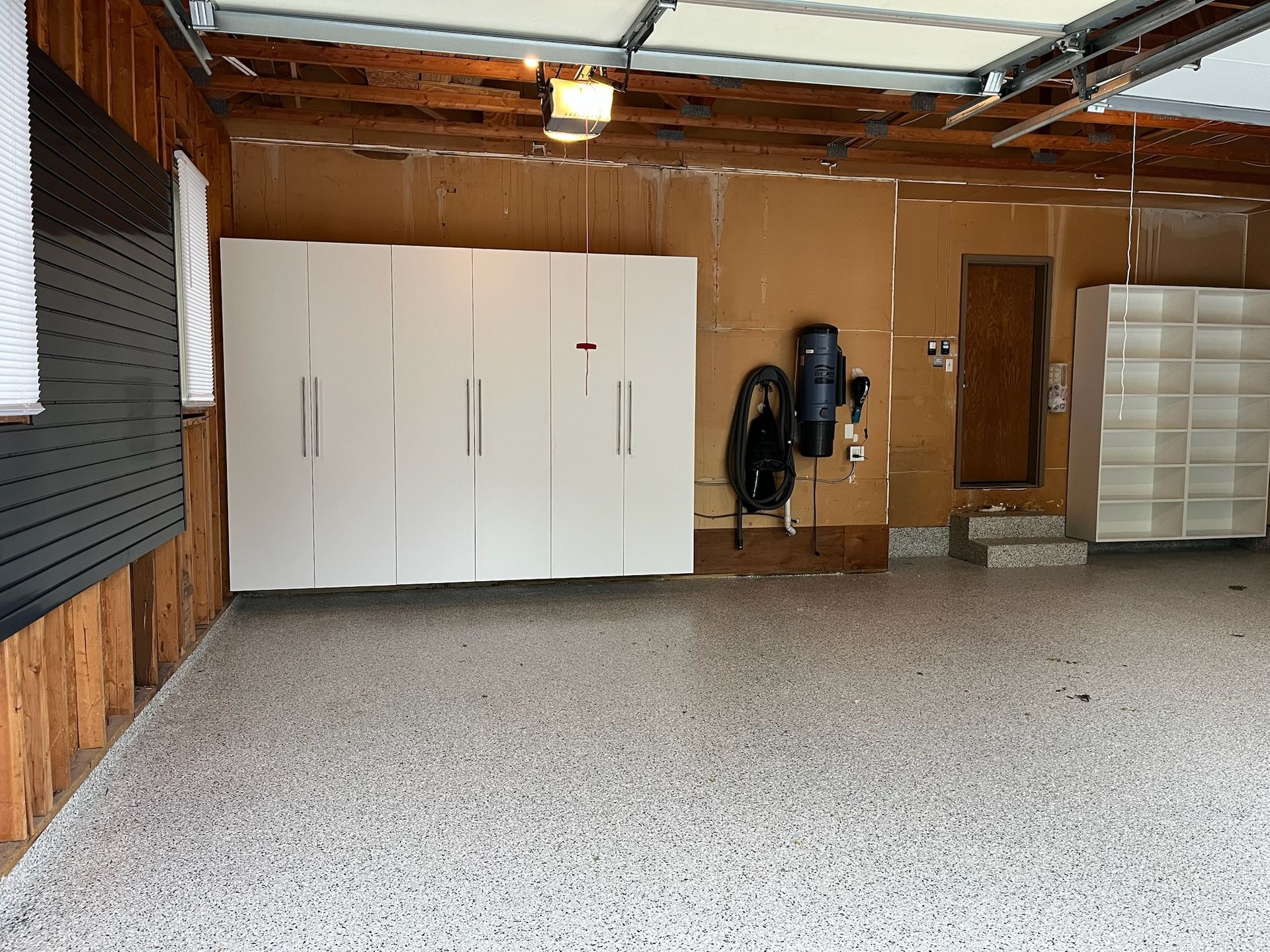 Empty garage with white cabinets, wooden walls, and a vacuum mounted near the back door.