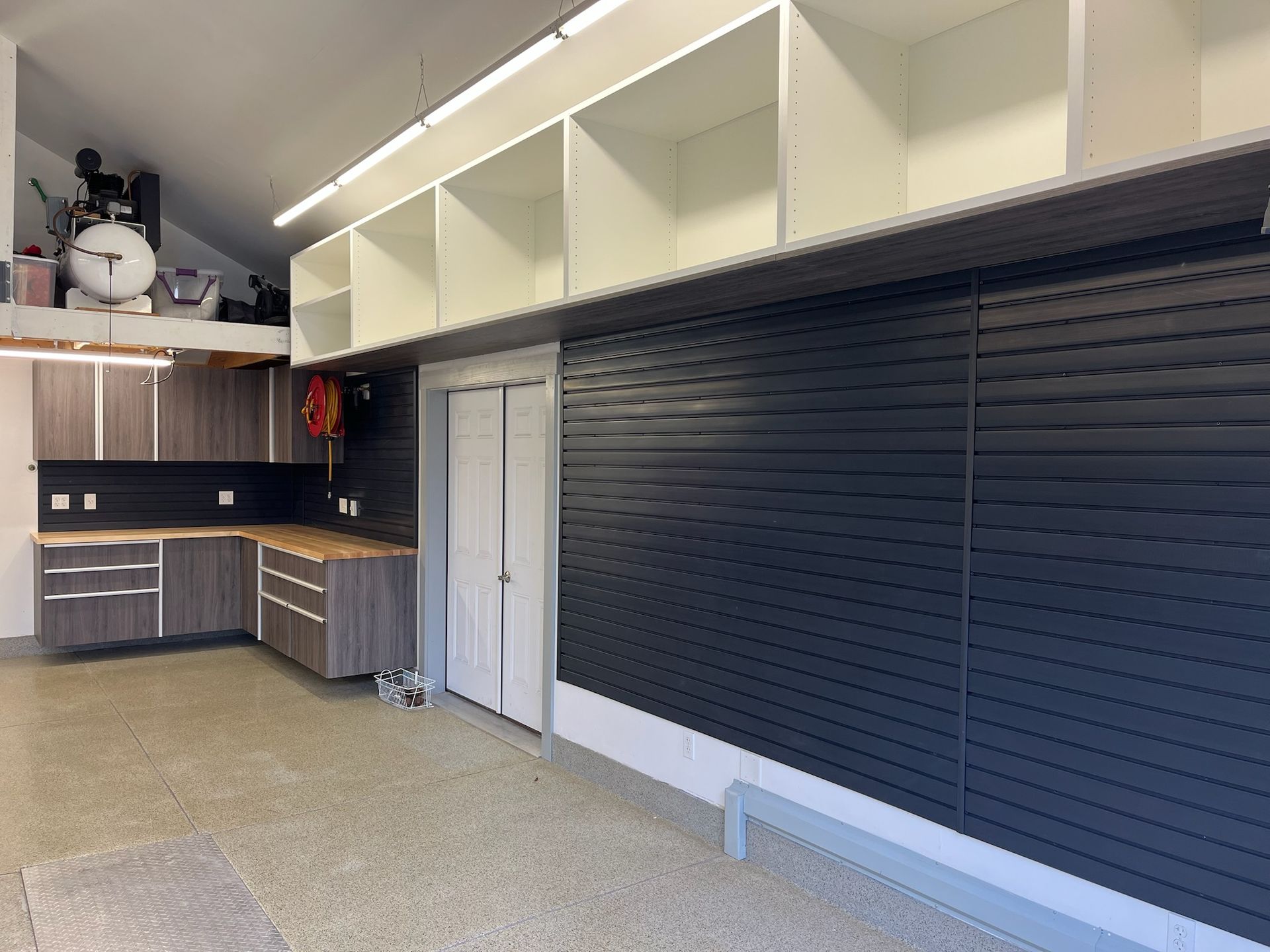 Garage workspace with gray cabinets, black slat wall, and white overhead storage shelves