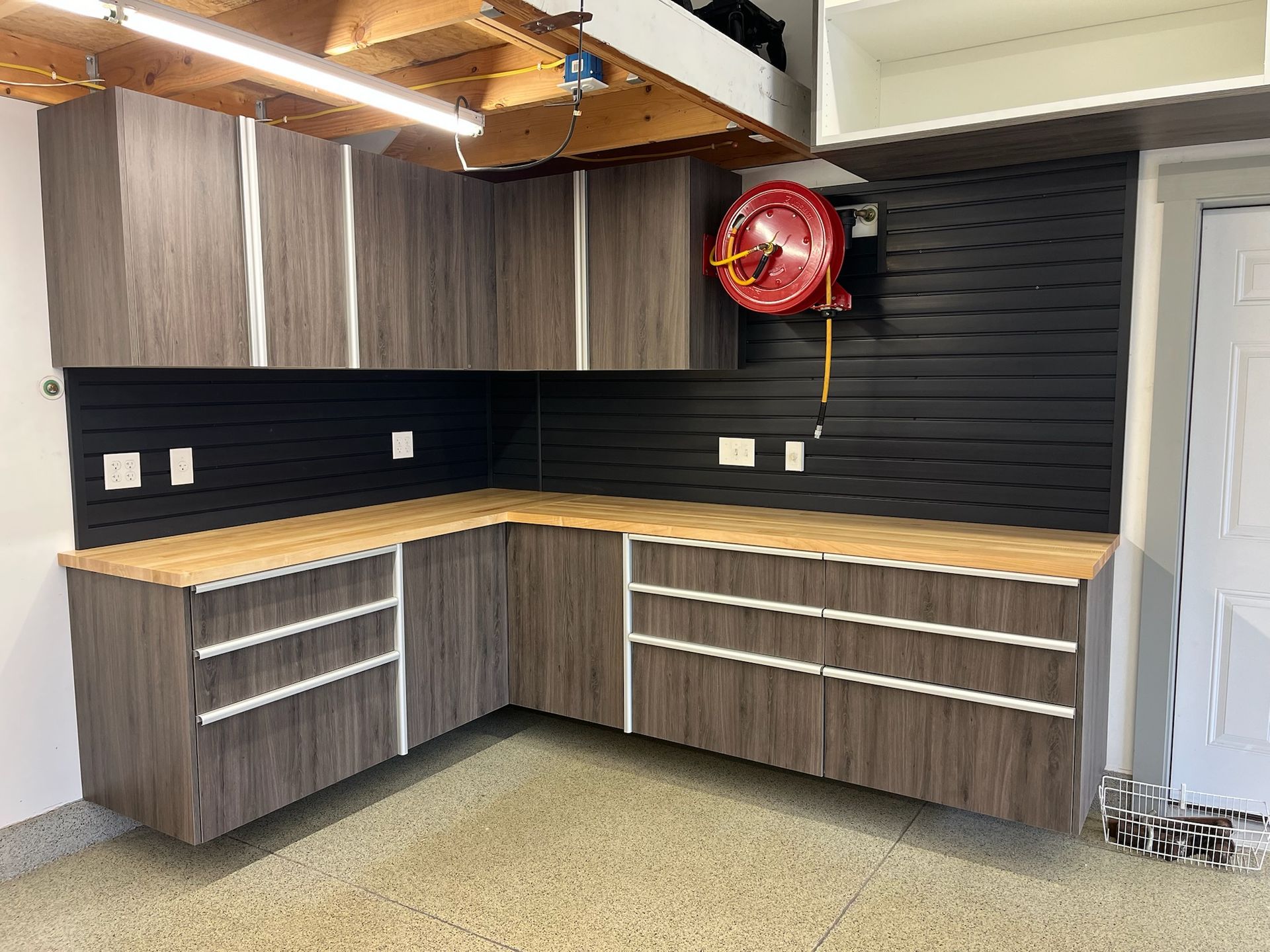 L-shaped garage cabinets with wood countertops and dark wood finish under a black slat wall.