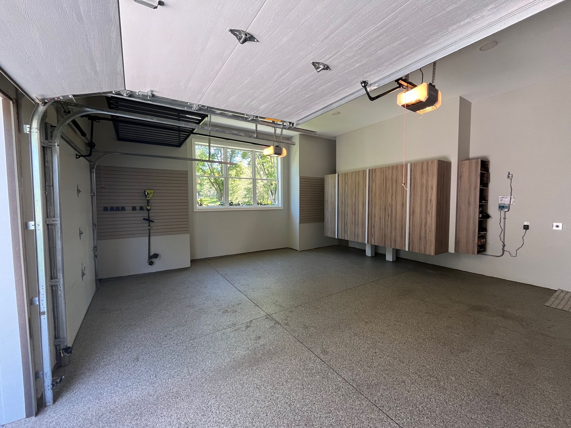 Empty garage with open door, speckled floor, window, wall cabinets, and overhead light.