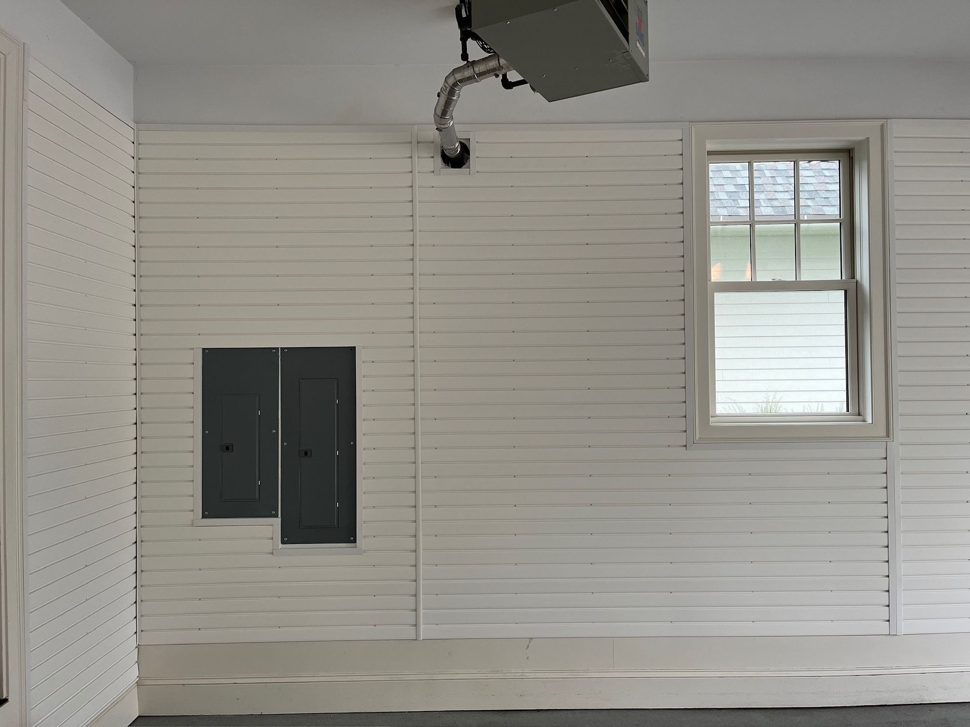 White paneled wall with a small window, electrical panels, and a garage door opener overhead.