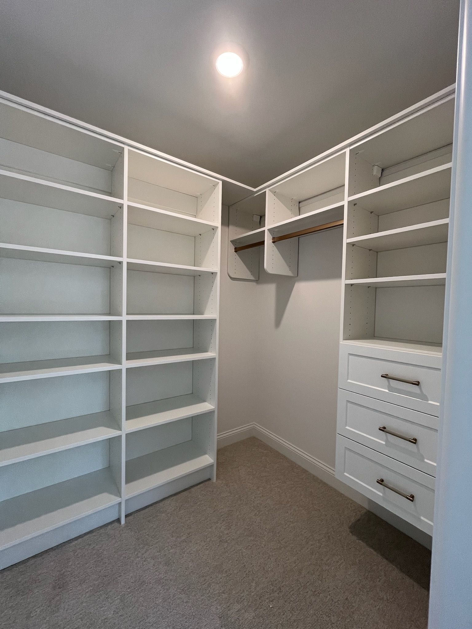 Walk-in closet with white shelving and drawers on both sides, gray carpet, and a ceiling light in the center.
