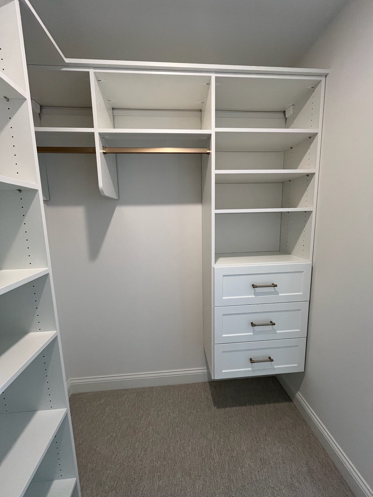 Empty walk-in closet with white shelving and drawers, beige walls, and gray carpet.