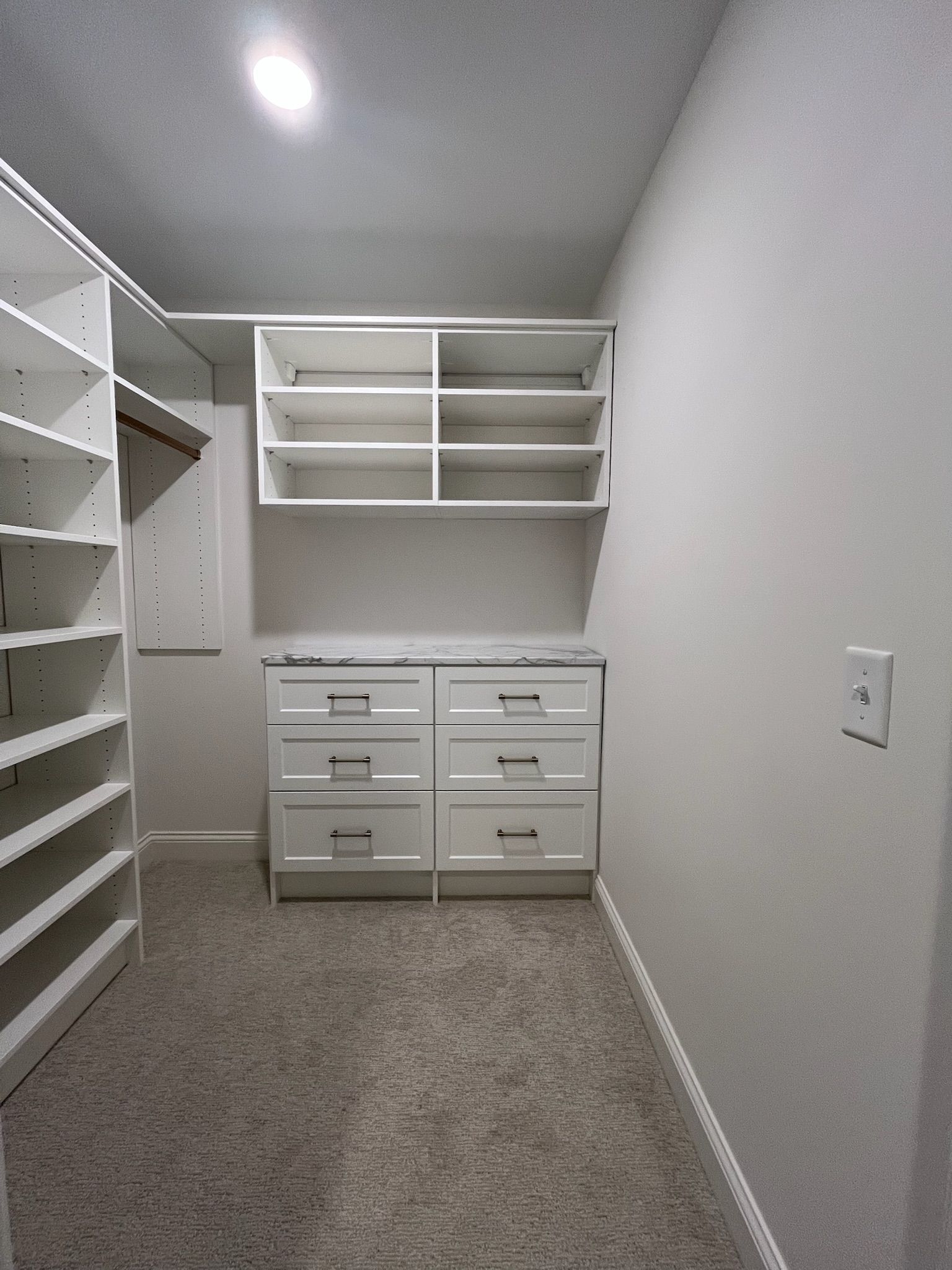 Empty walk-in closet with white shelves and drawers, carpeted floor, and sloped ceiling.