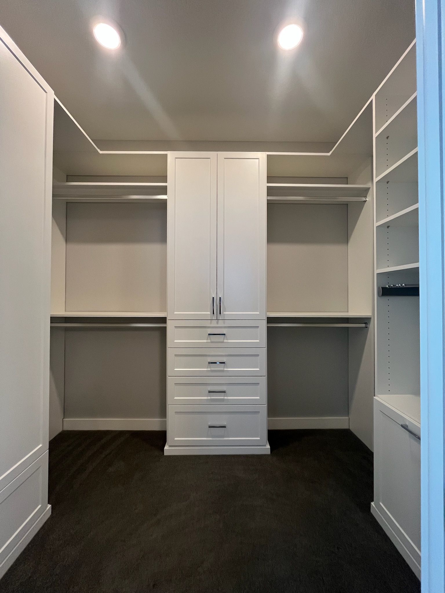 Empty walk-in closet with white built-in shelves, hanging rods, and drawers on dark flooring.