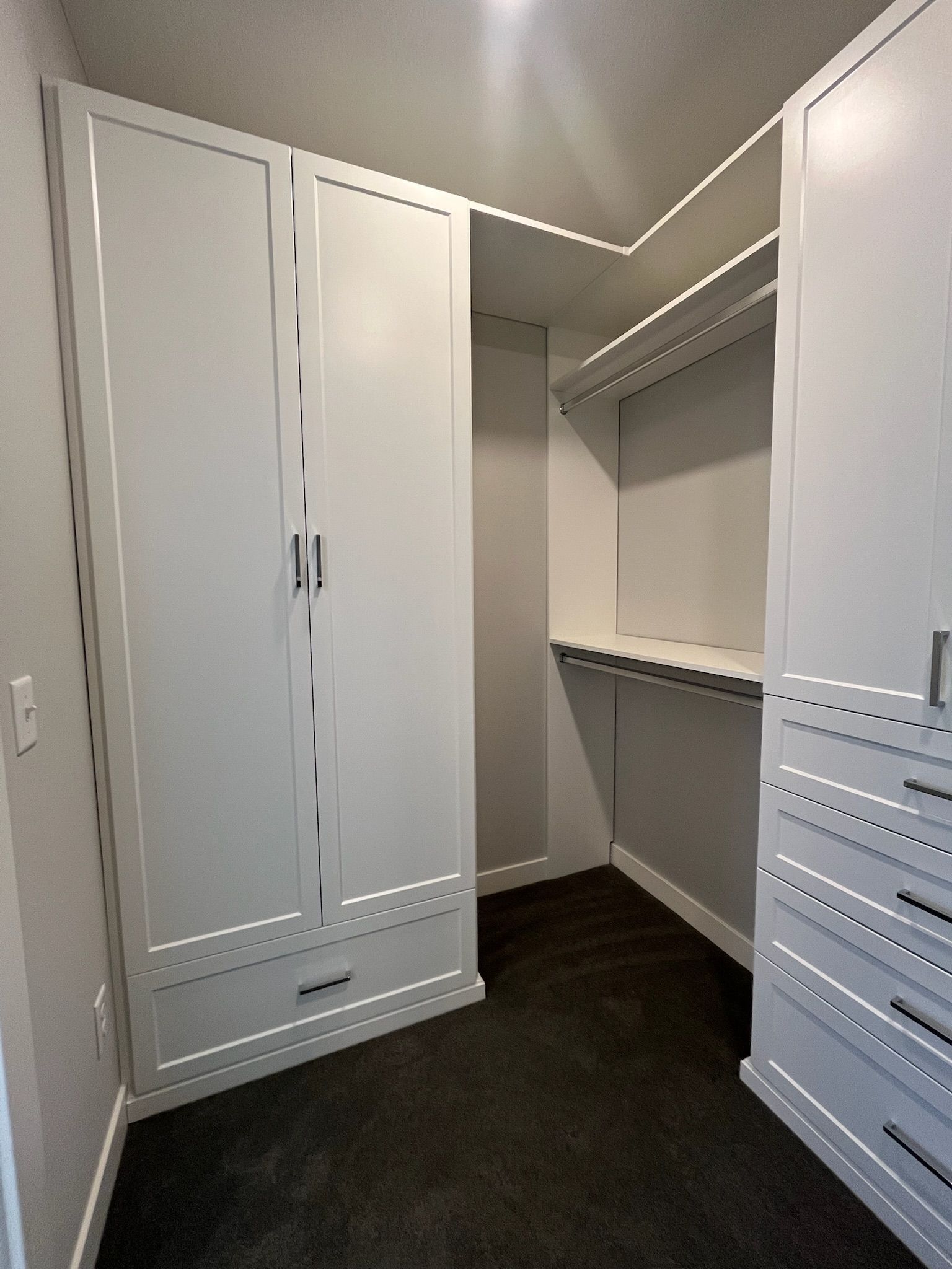 Empty walk-in closet with white cabinets, hanging rods, and dark carpet flooring.
