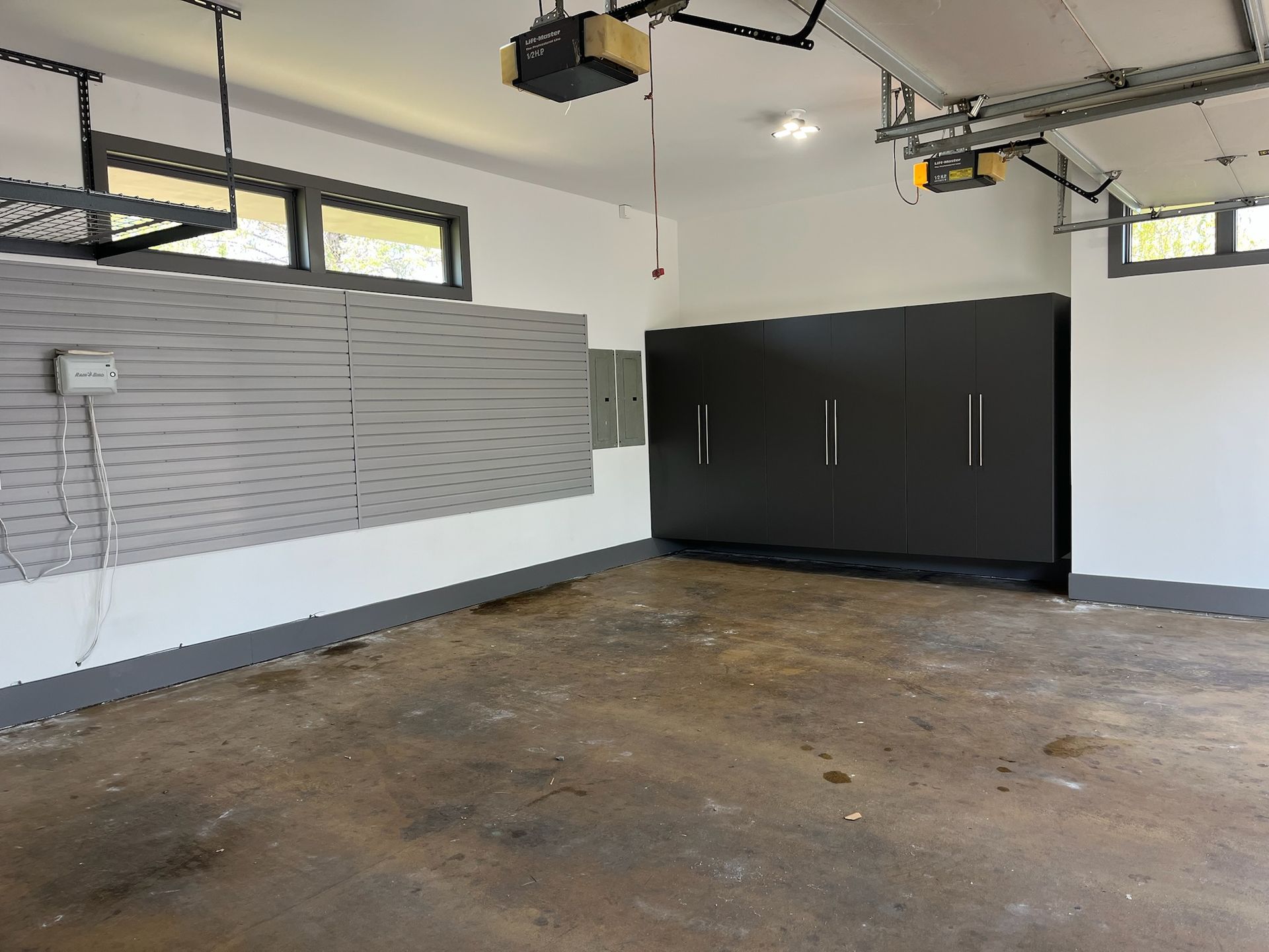 Empty garage with a concrete floor, white walls, black storage cabinets, and a row of small windows.