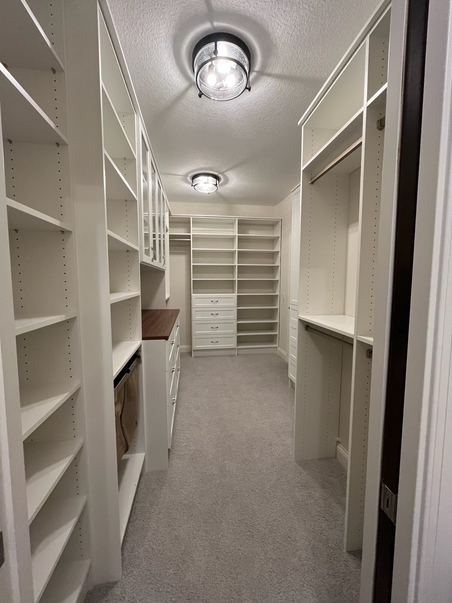 Narrow white walk-in closet with shelves, hanging rods, and carpeted floor under ceiling lights
