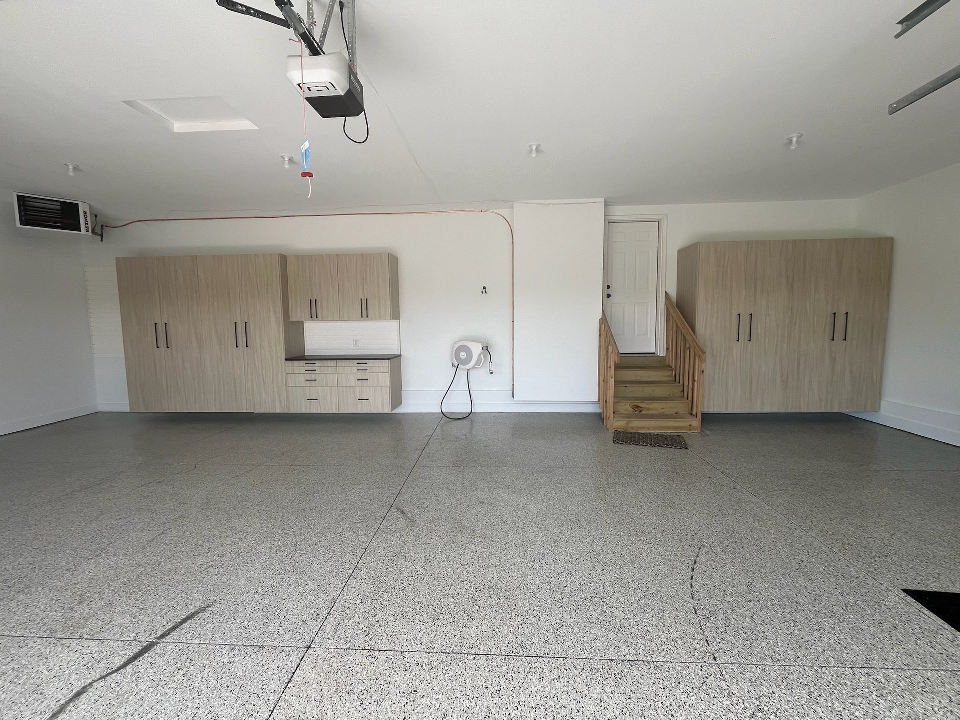 Empty garage with gray speckled floor, white walls, cabinets, and a wooden staircase at the back.