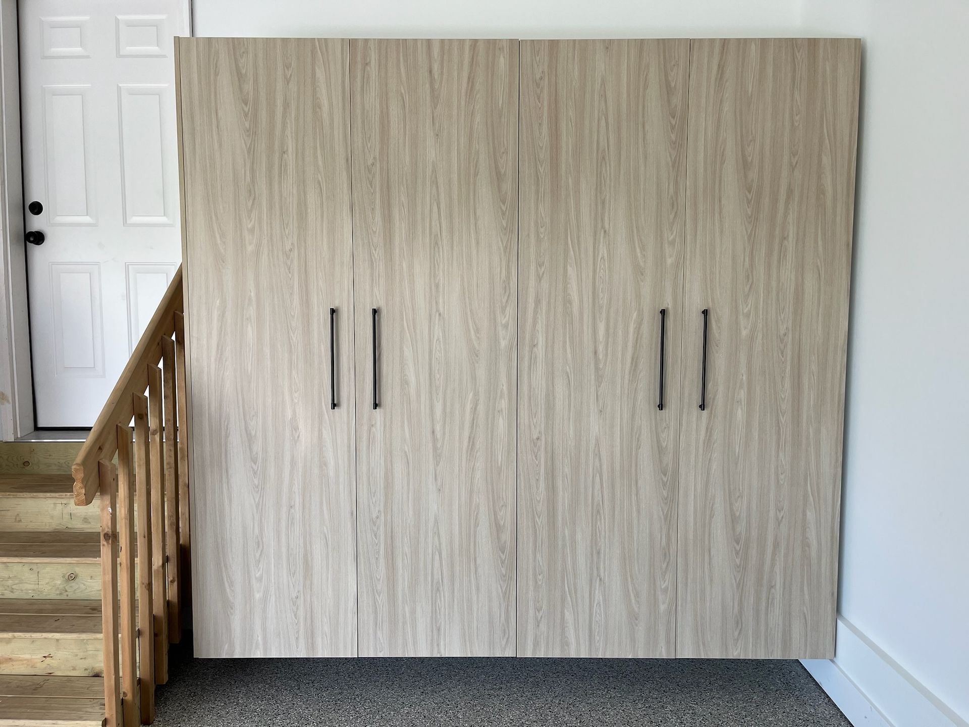 Light wood wall cabinet with four handles beside a staircase and white door.