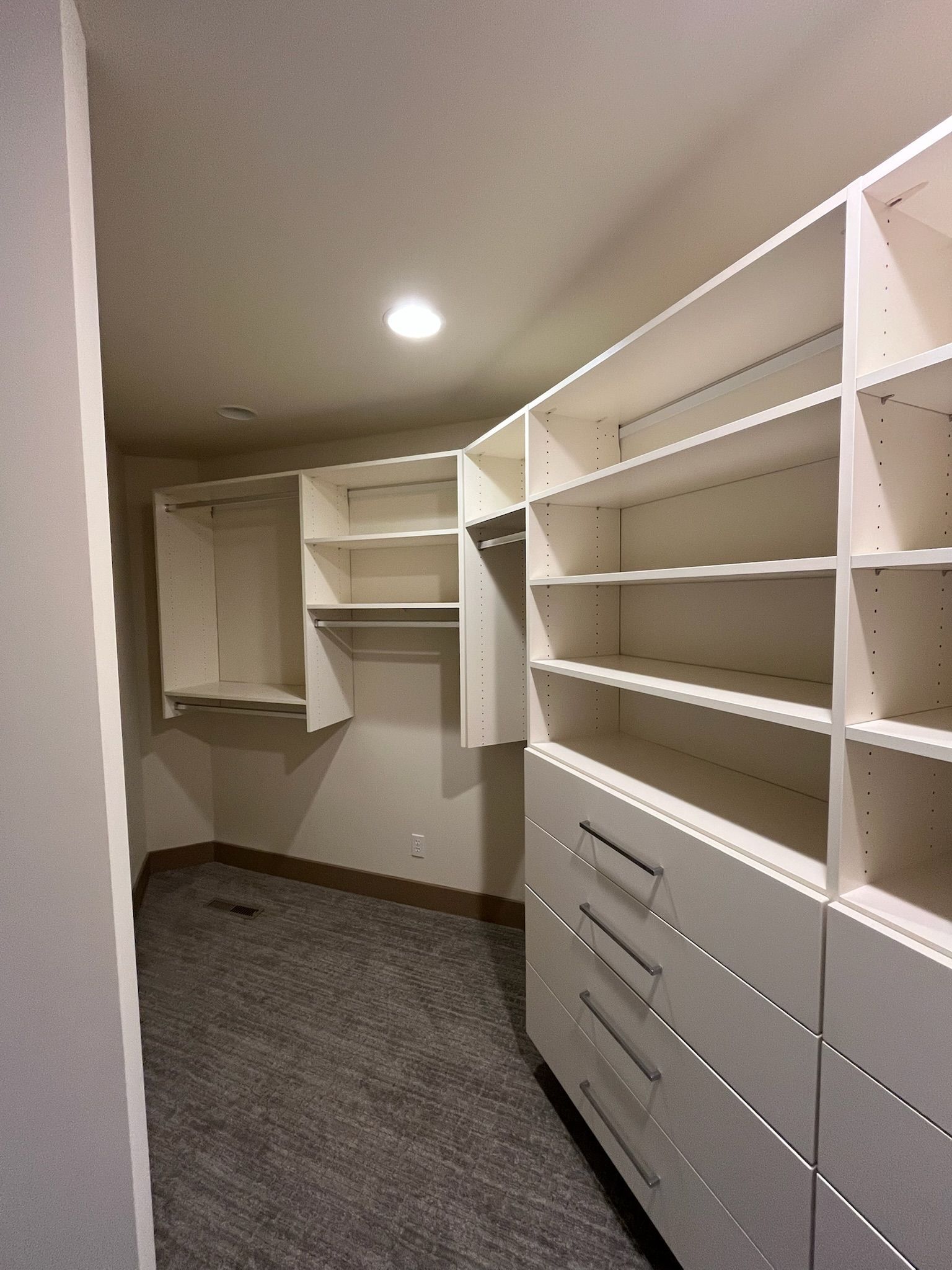 Empty walk-in closet with white built-in shelves and drawers, gray carpet, and a ceiling light