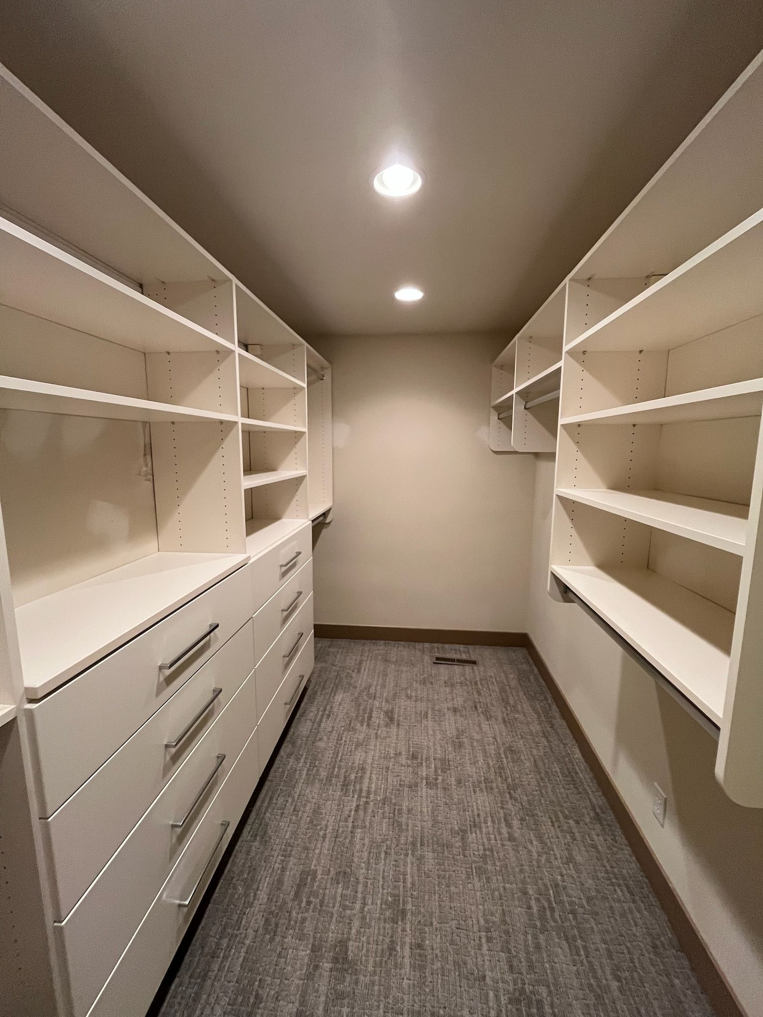 Narrow walk-in closet with white shelves and drawers, carpeted floor, and recessed ceiling lights.