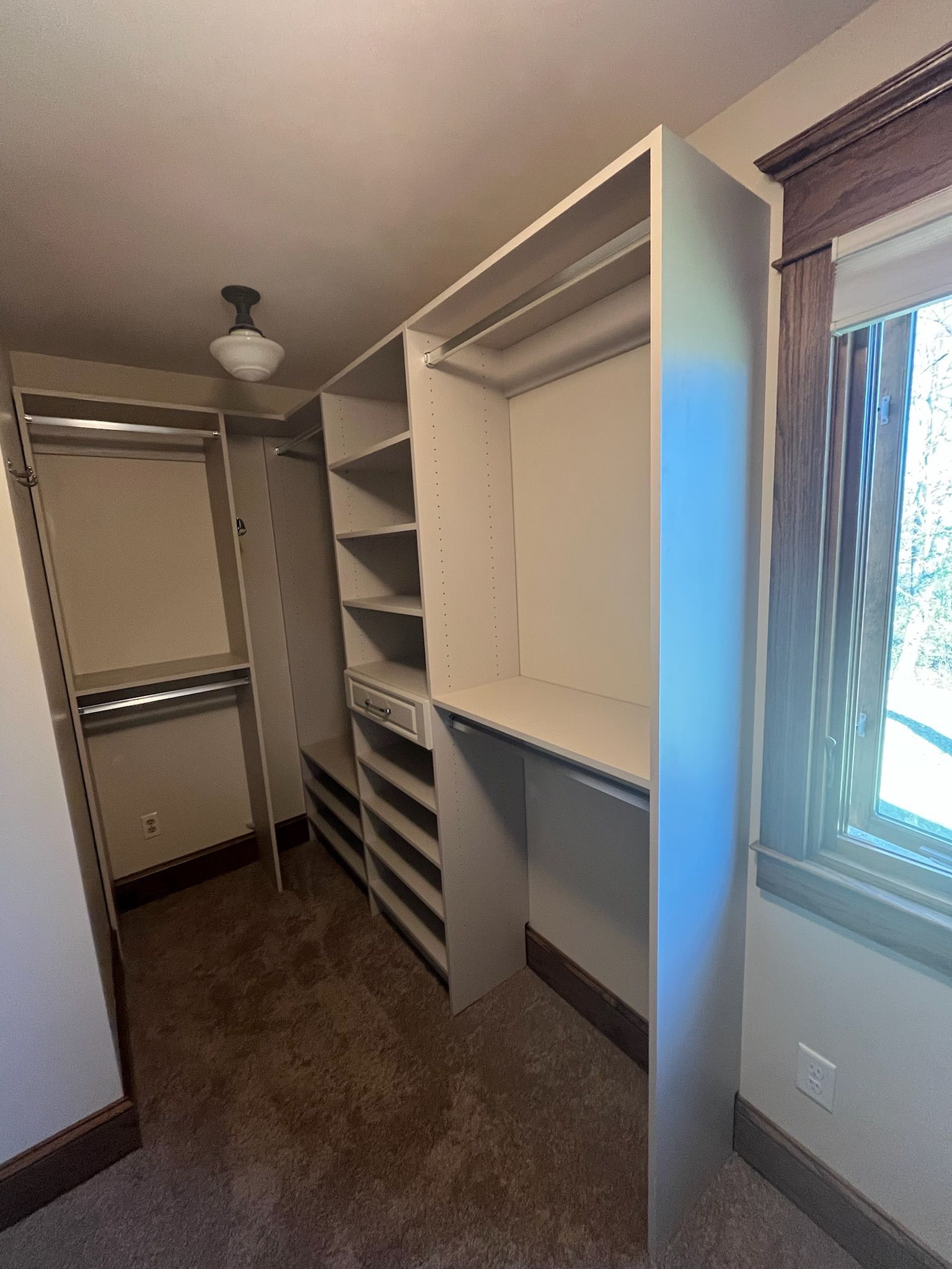 Walk-in closet with white shelving and hanging rods beside a window with curtains.