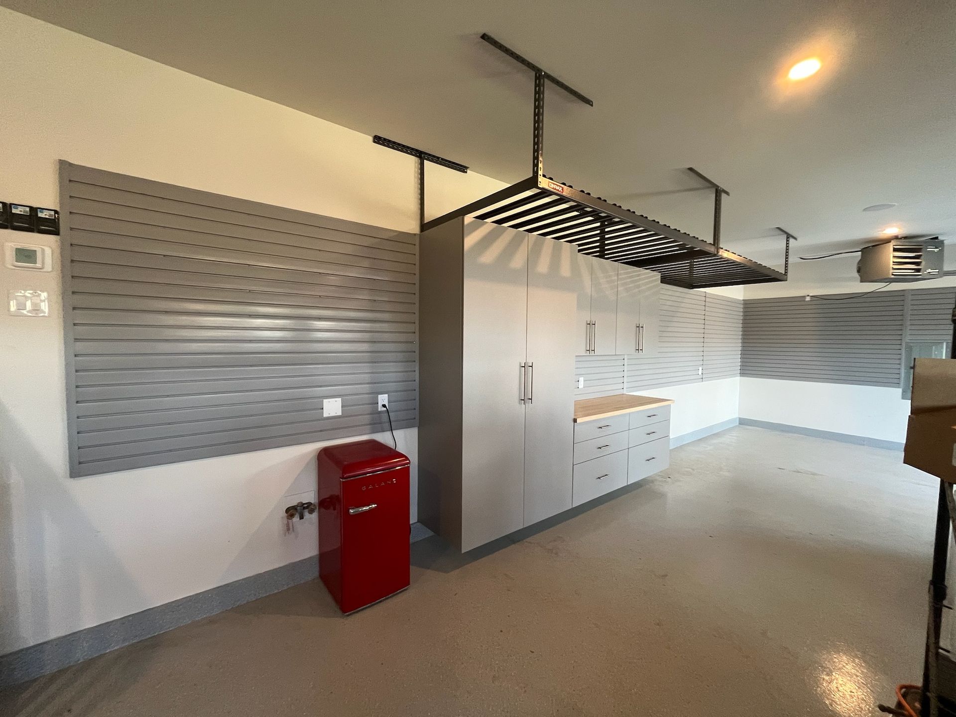 Modern office kitchen with white counters, grey walls, hanging racks, and a red bin.