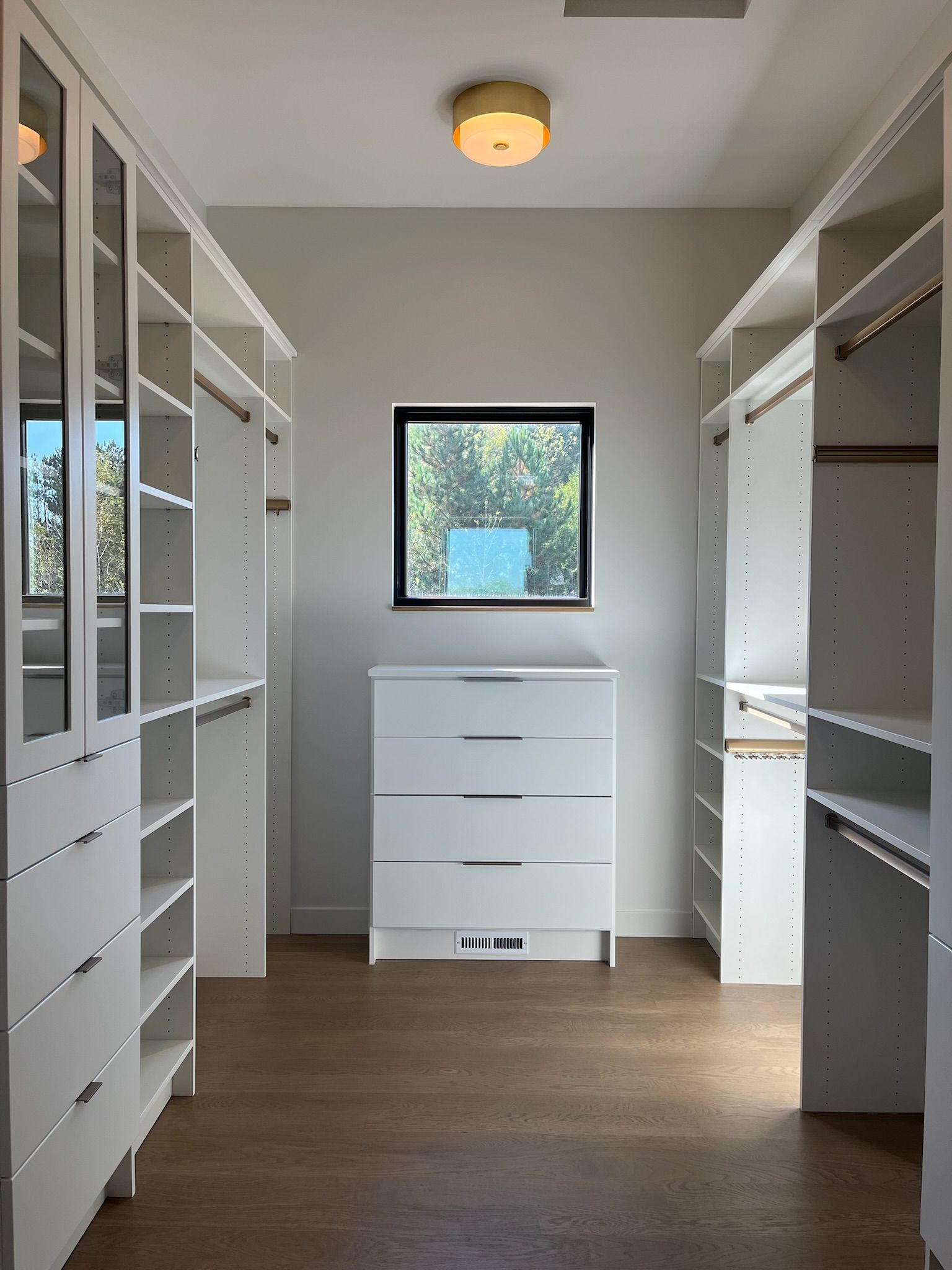 Empty walk-in closet with white shelves, a central dresser, and a small window.