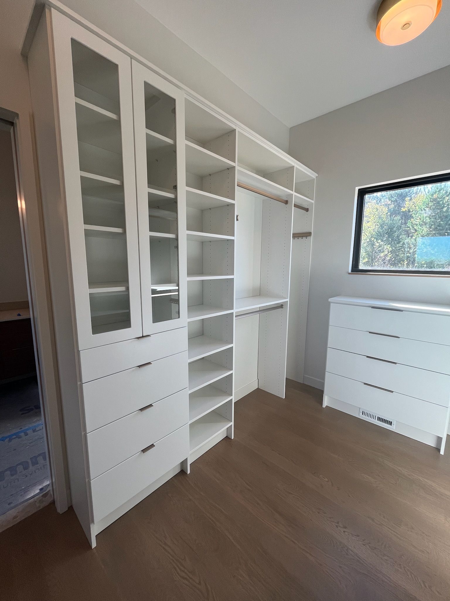White walk-in closet with built-in shelves, drawers, and hanging space, plus a matching dresser by a window.