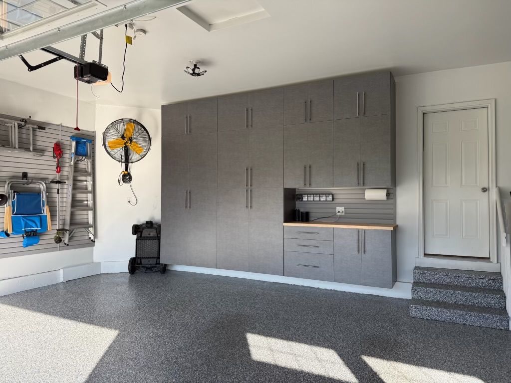 Garage interior with gray cabinets, wall fan, storage shelves, and sunlight on carpeted floor