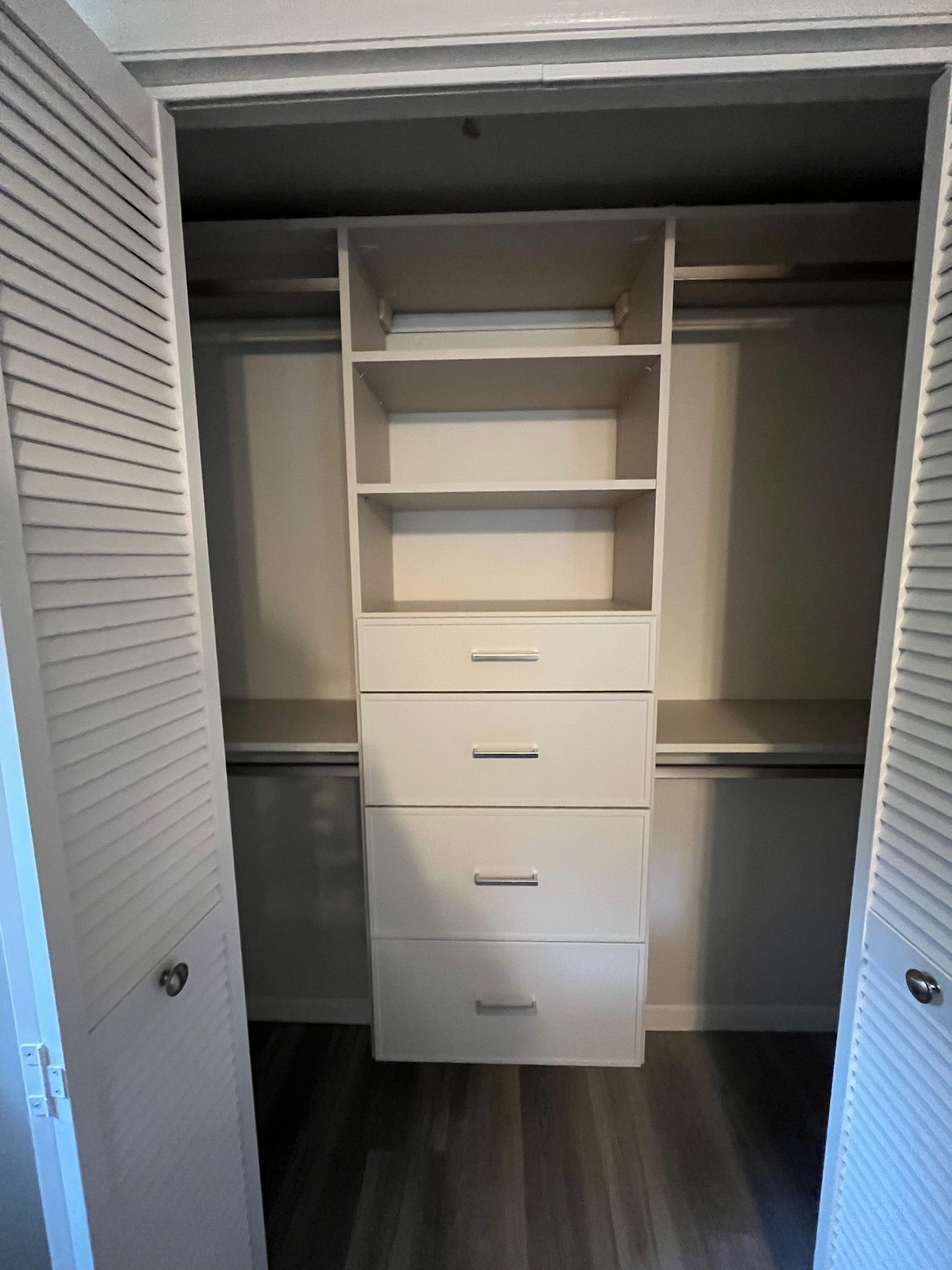 Open closet with white shelves and drawers, flanked by louvered doors on dark flooring.