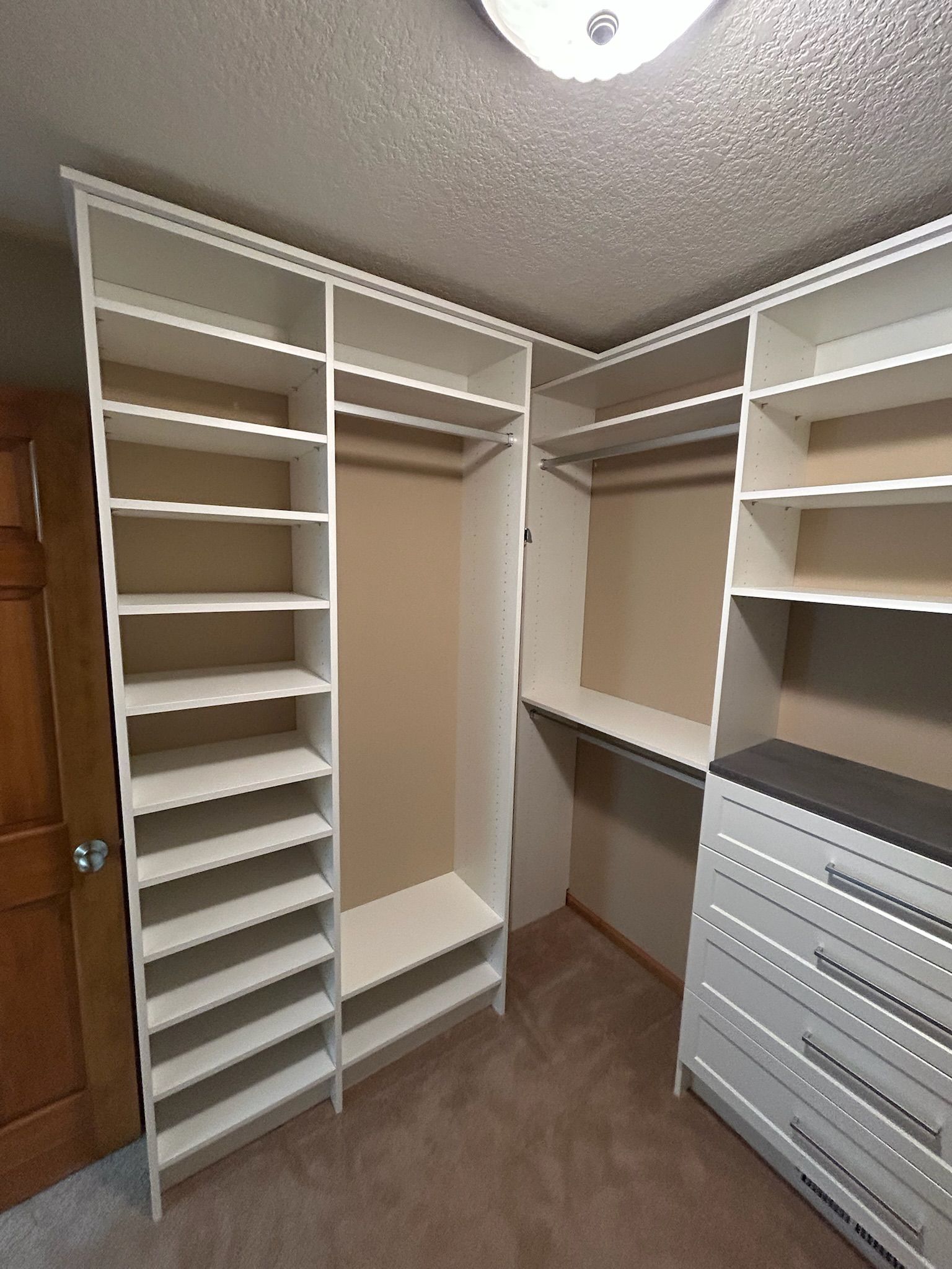 Walk-in closet with white shelving, hanging rods, and a gray drawer unit on brown carpet.