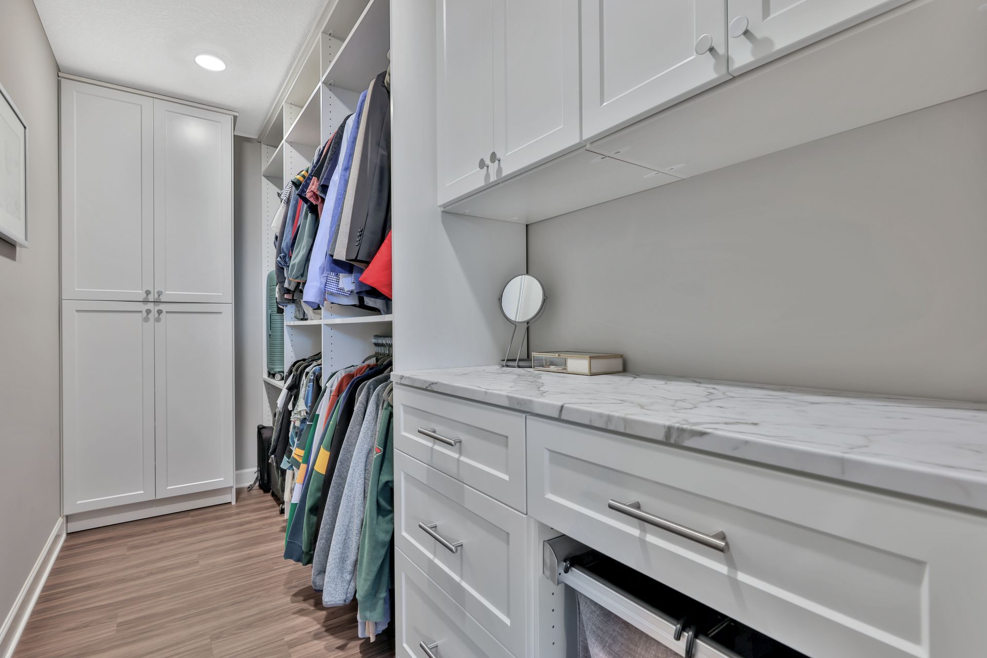 Built-in closet with white cabinets, hanging clothes, and a countertop mirror over a wood floor.